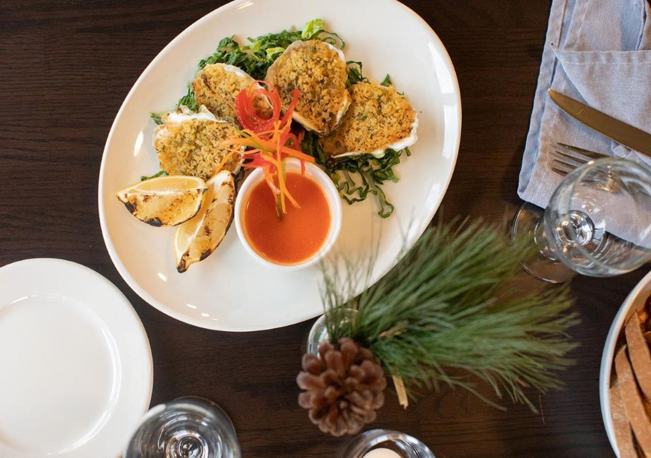 A platter of baked oysters topped with breadcrumbs, served with dipping sauce, lemon wedges, and greens.