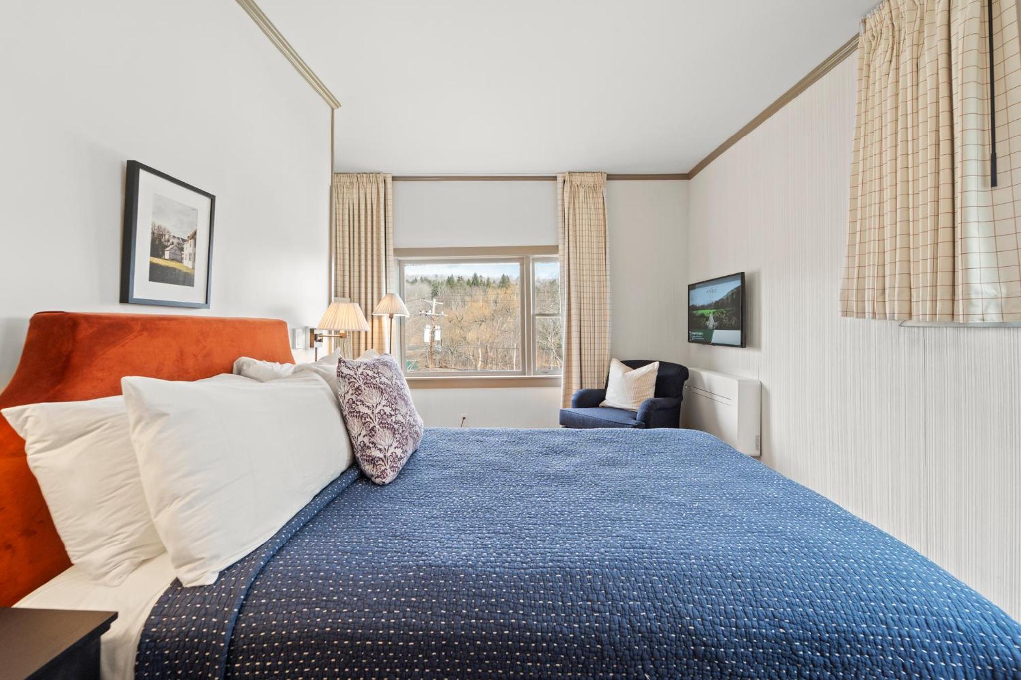 A bedroom with an orange upholstered headboard, blue quilt, corner armchair, and window looking out to trees.
