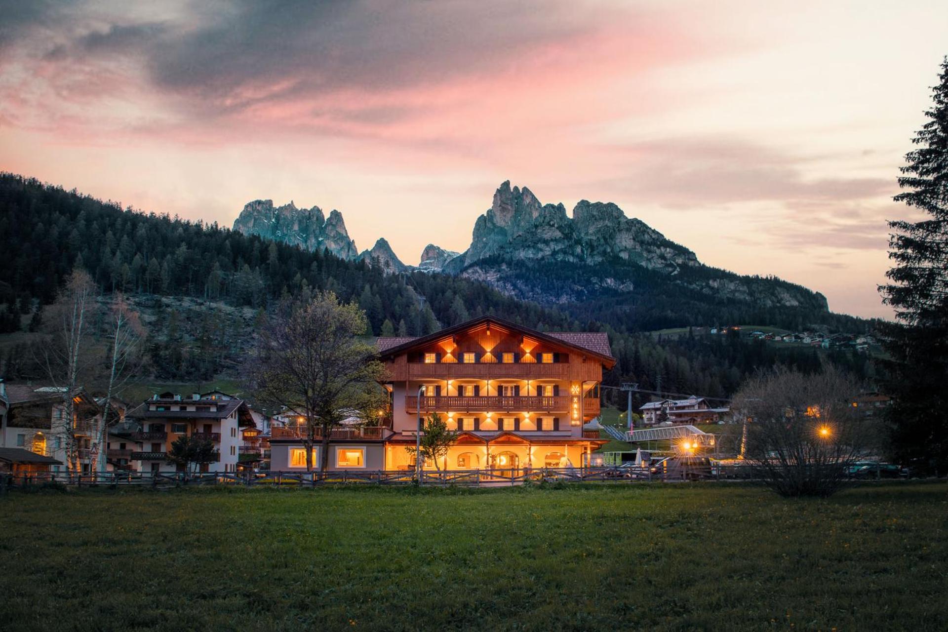 Illuminated facade of X Alp Hotel at dusk with towering alpine peaks and pine forests in the background.