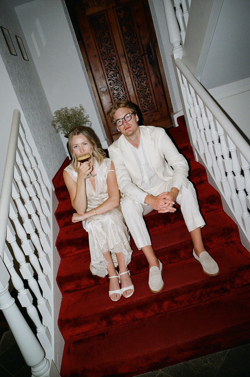 Perrie and Walt sitting on a red-carpeted staircase, Perrie mid-sip from a cocktail glass