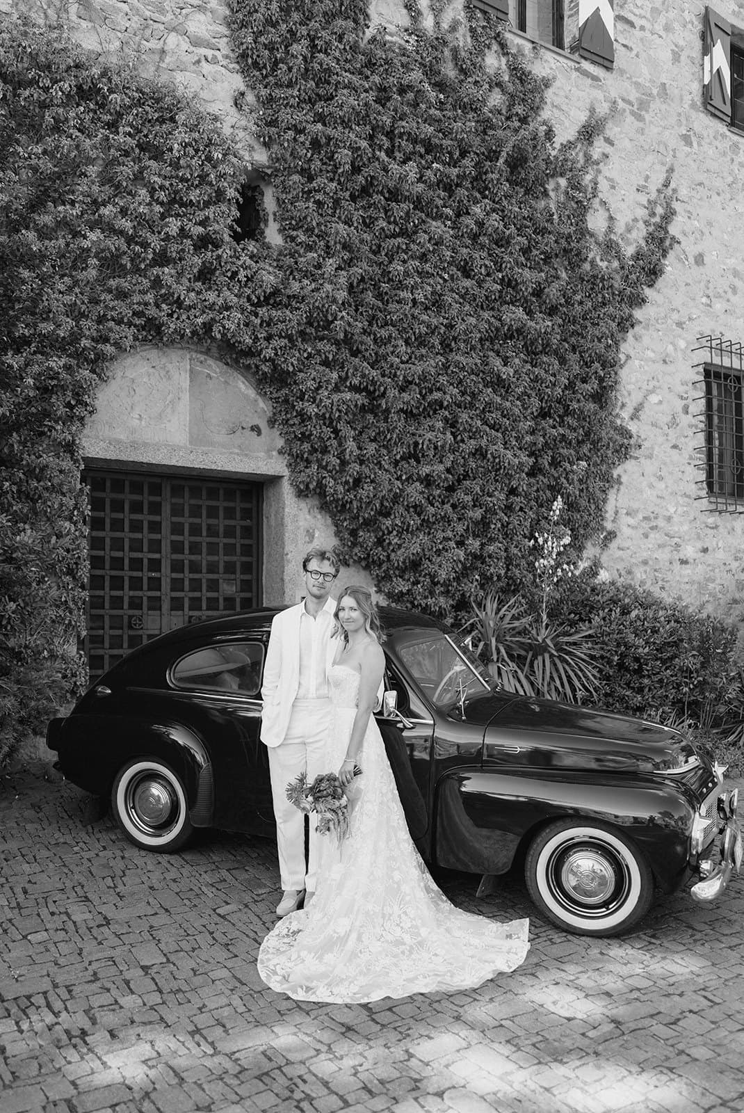 Walt and Perrie beside a vintage car in front of an ivy-covered castle wall, in black and white