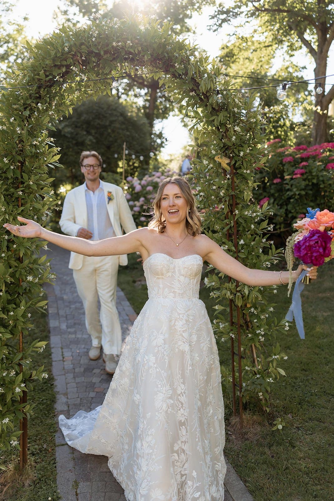 Perrie with her arms open and a bright bouquet in one hand, walking out of a garden arch with Walt behind her