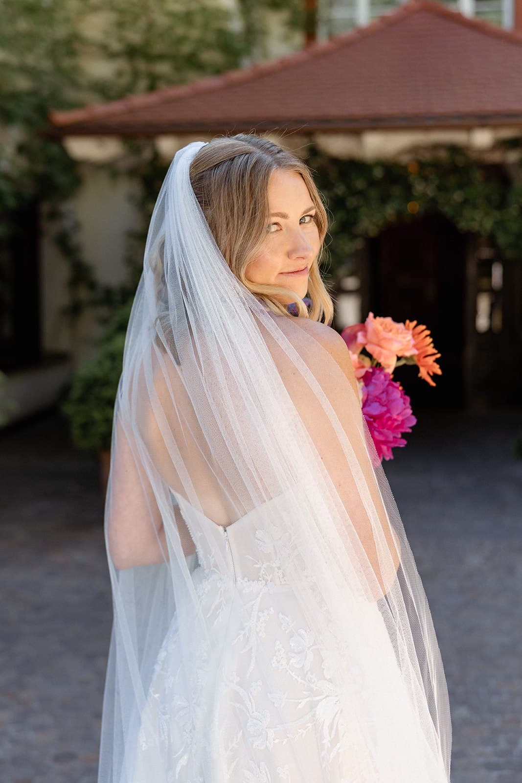 Perrie in her veil at Castel Fragsburg, looking back over her shoulder with a bouquet of pink and orange flowers