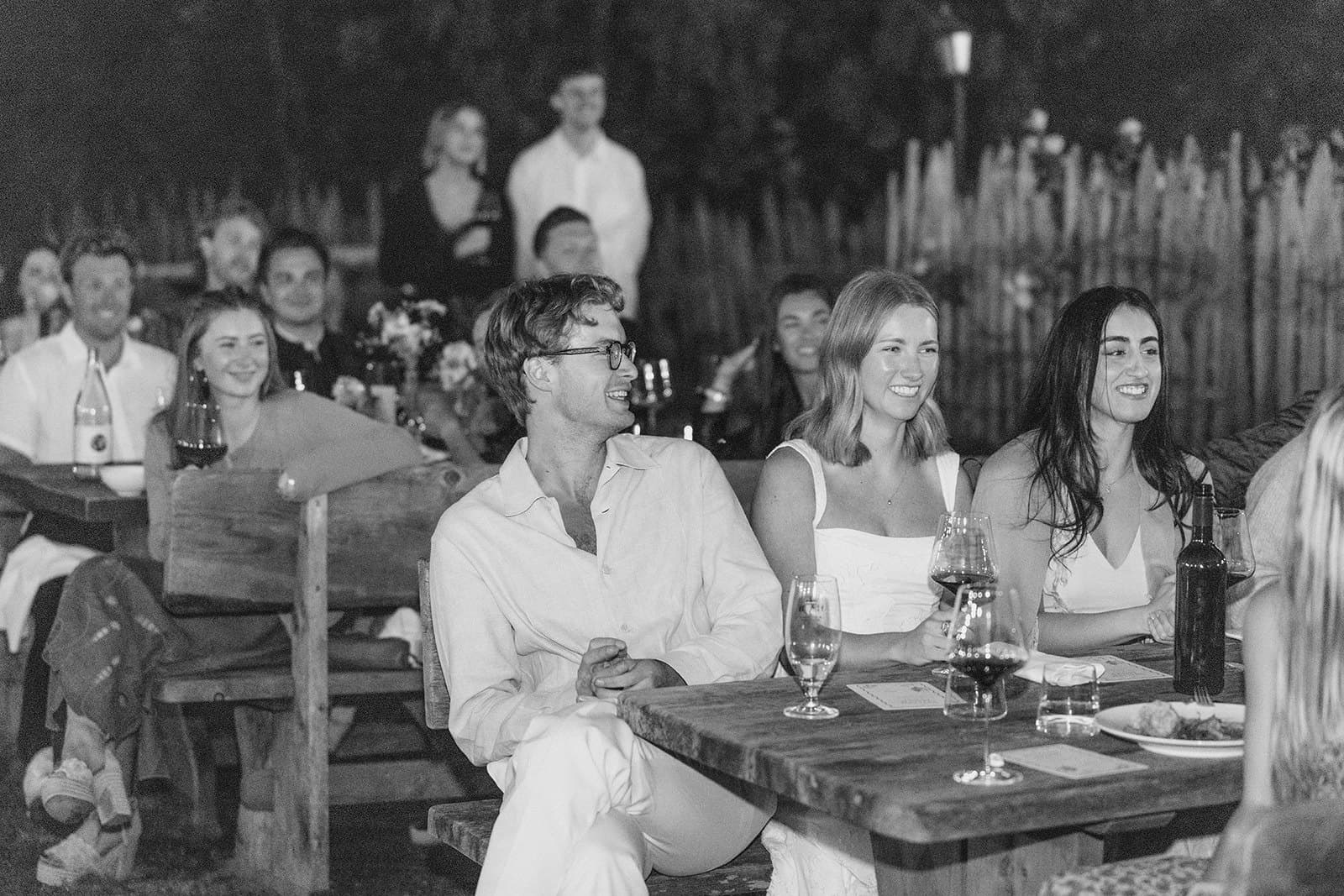 Guests laughing at long wooden tables during the welcome dinner, in black and white