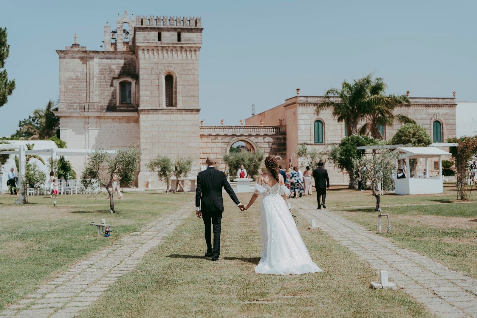 A bride and groom walk away from the camera on a stone path through a grassy courtyard.