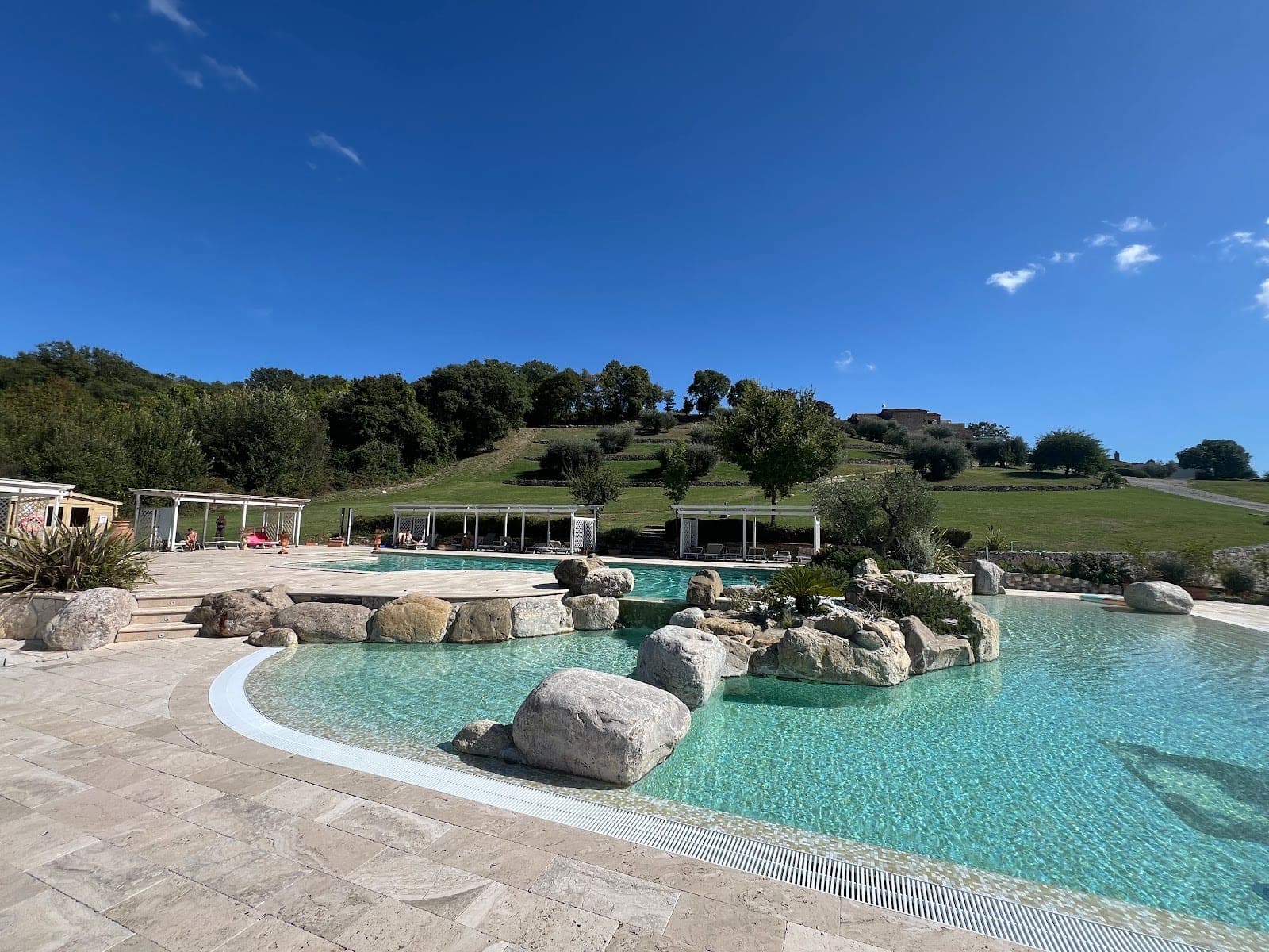 A large, clear swimming pool with rock features and surrounding paved area, set in a green landscape under a blue sky.