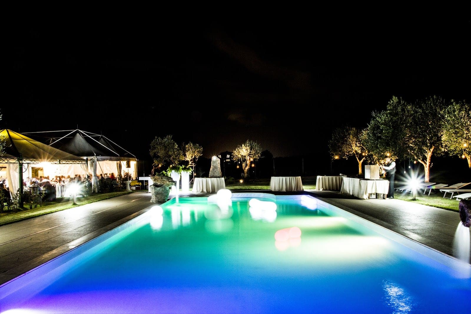 A swimming pool with colored lights at night, with reception tents in the background.