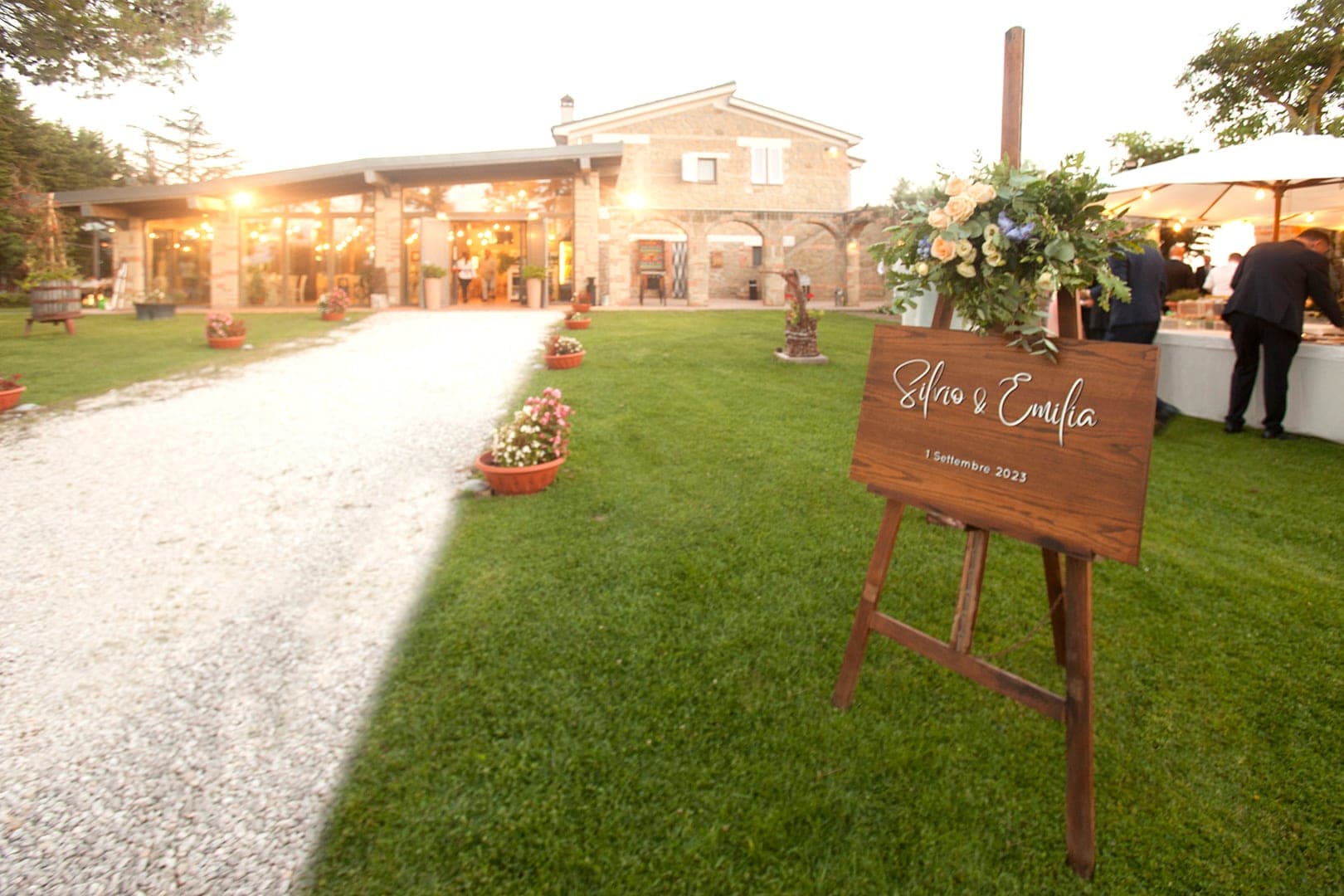 A wooden welcome sign with names and a date stands on the lawn in front of a building.