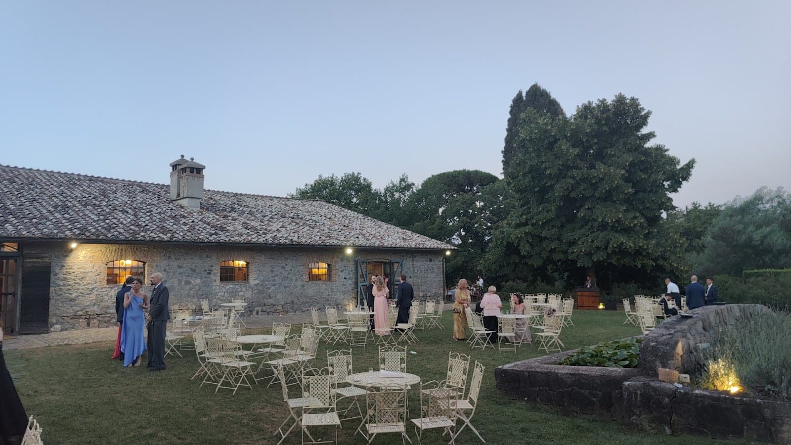 Wedding reception setup with tables and chairs on a lawn in front of a stone building.