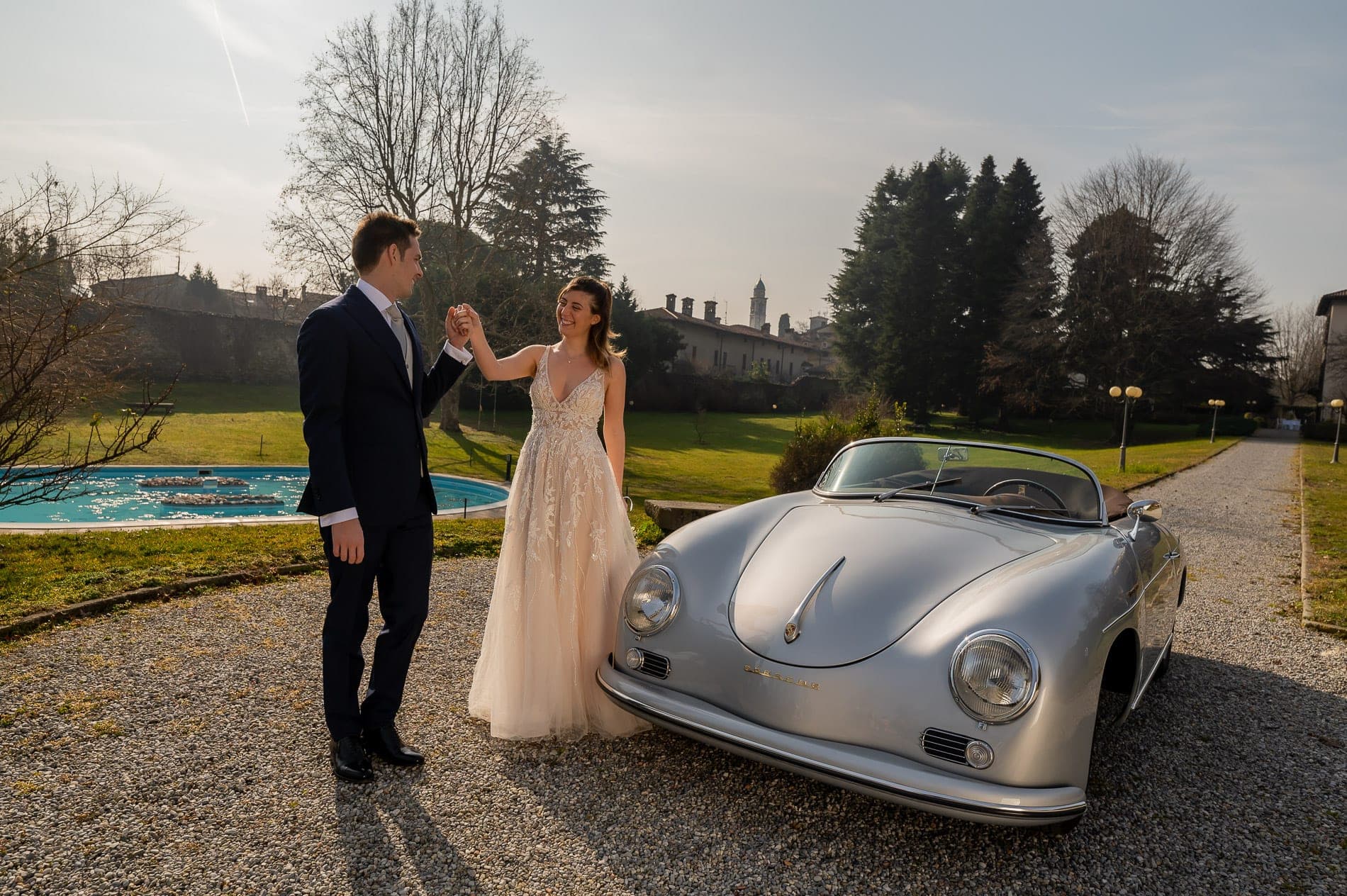 Couple in wedding attire with a vintage car on the grounds of a wedding venue.