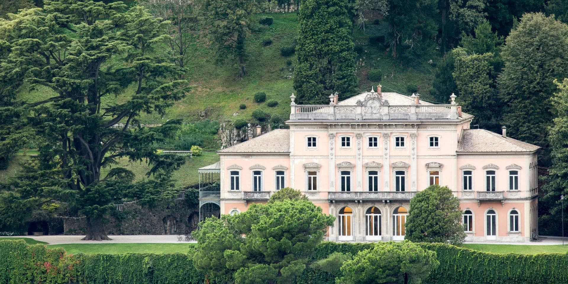 A grand pink villa with white trim sits behind manicured hedges and lush trees.