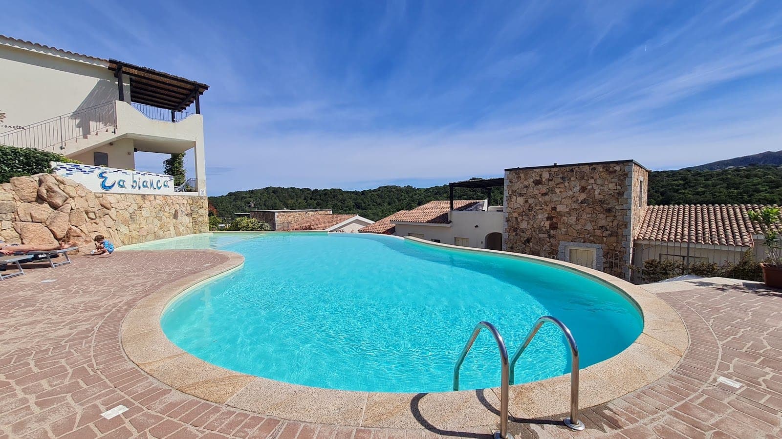 A swimming pool with blue water and a paved area around it, with buildings and hills in the background.