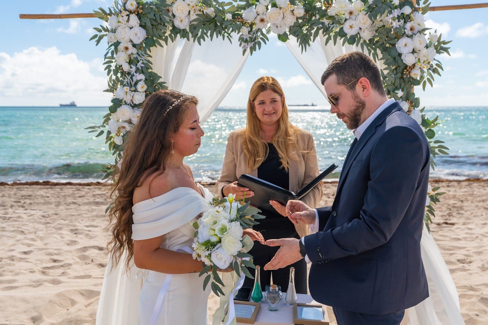 A wedding ceremony takes place on a sandy beach under a floral arch with the ocean behind.