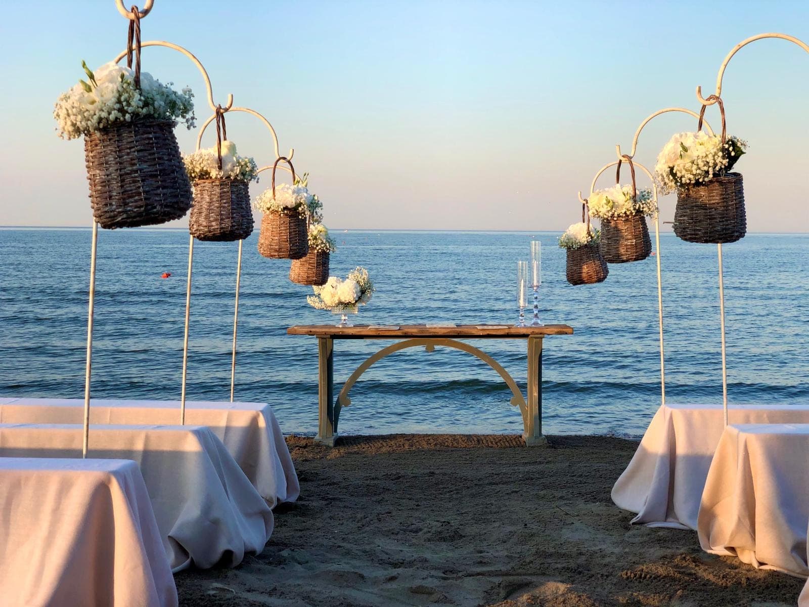 Beach ceremony setup with hanging flower baskets and tables by the sea.