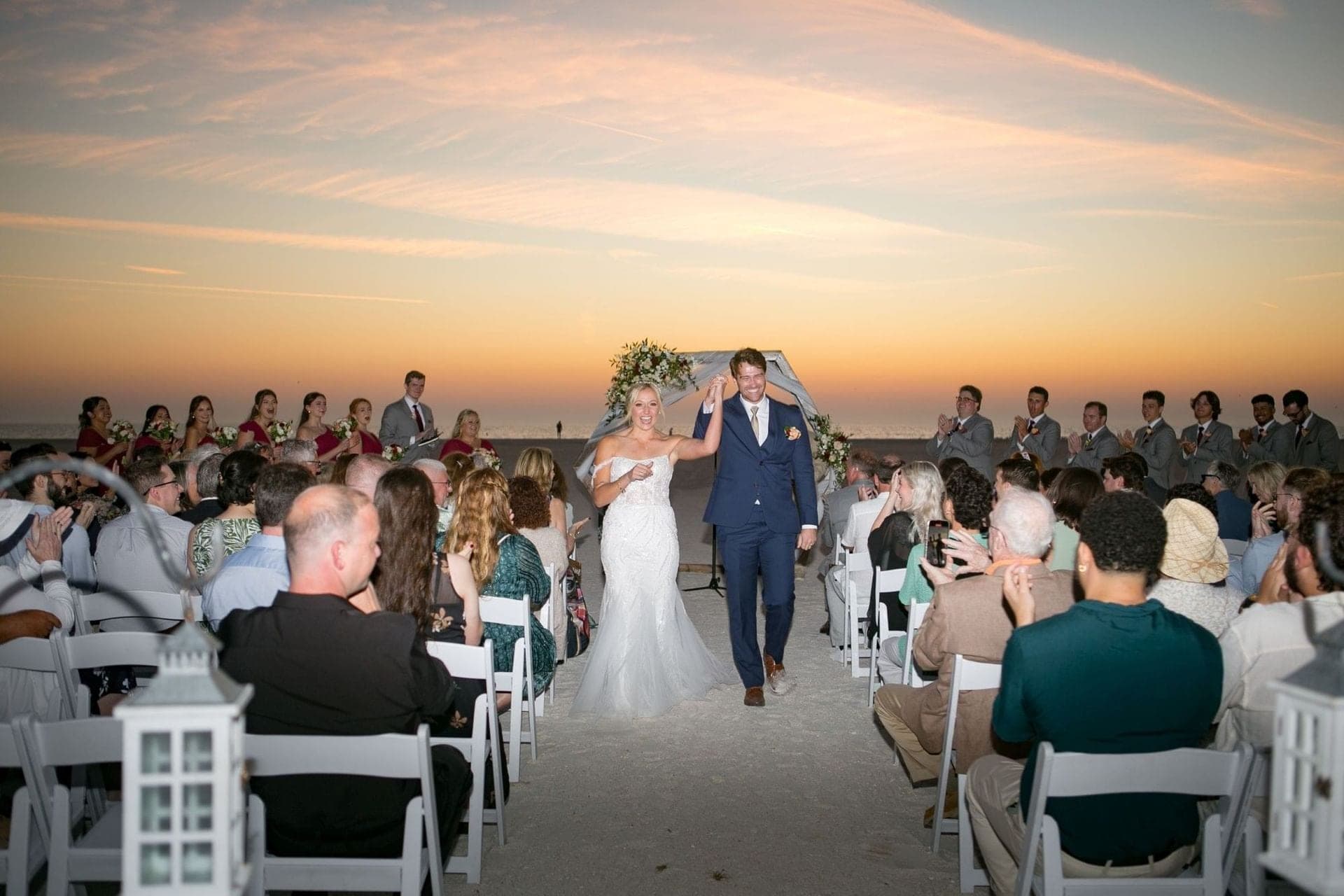 Bride and groom walk down the aisle on a beach at sunset.