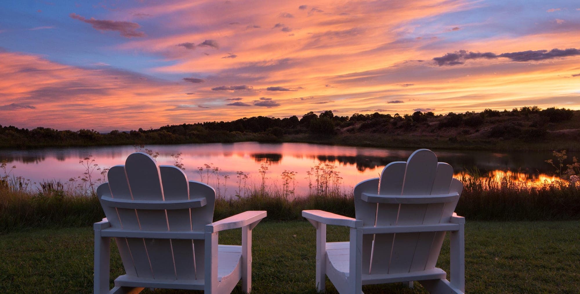 Two white Adirondack chairs facing a lake at sunset with a colorful sky.