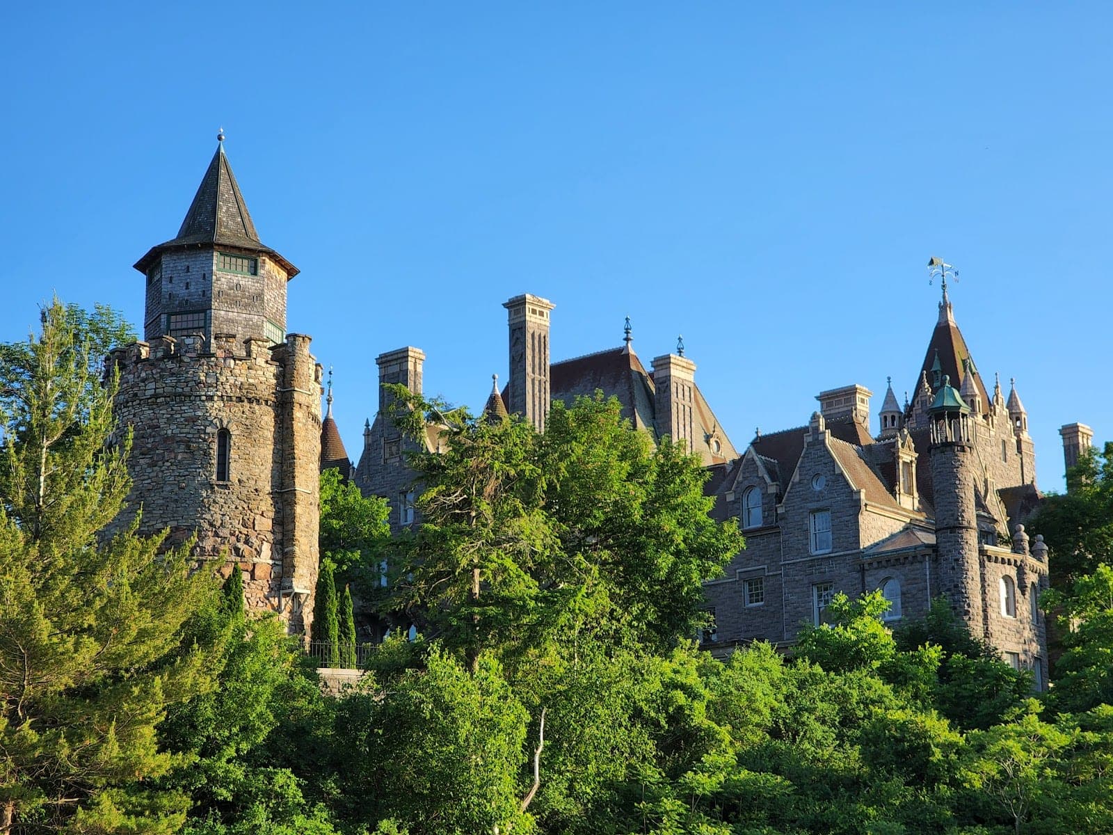 A stone castle with multiple towers and spires surrounded by lush green trees under a clear blue sky.