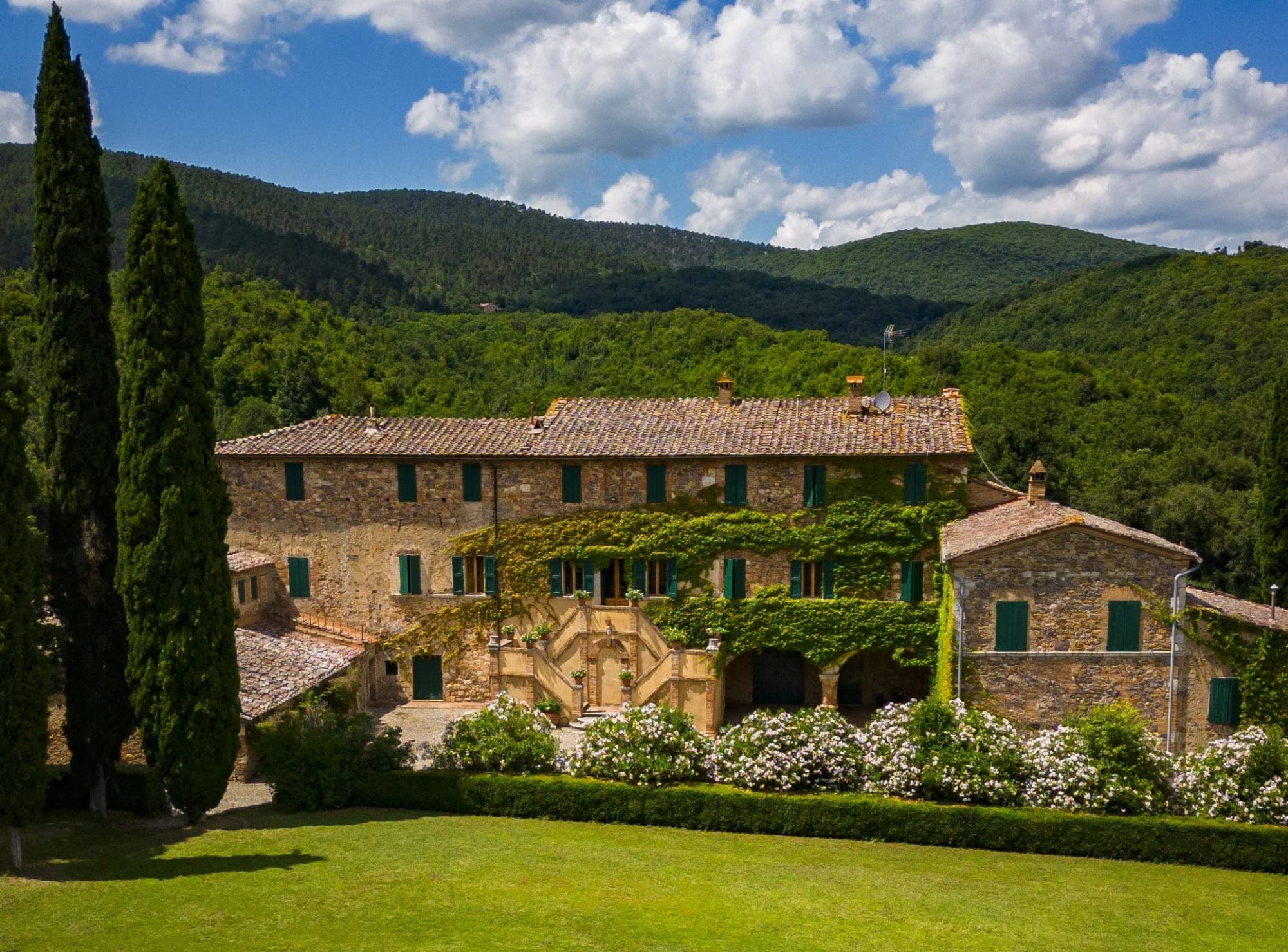 Stone building with green shutters and ivy, set against a backdrop of green hills under a blue sky with clouds.