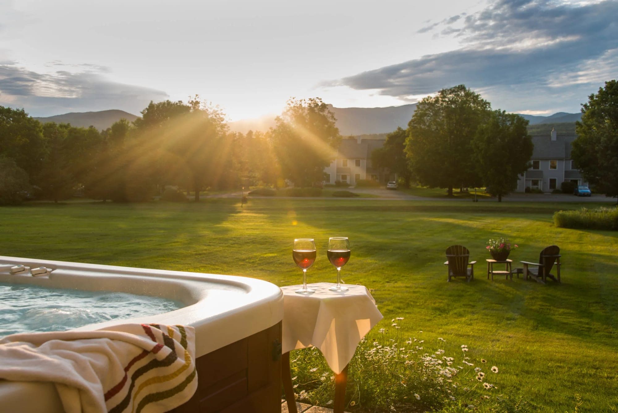 Hot tub and wine glasses on a table with a lawn and buildings at sunset.