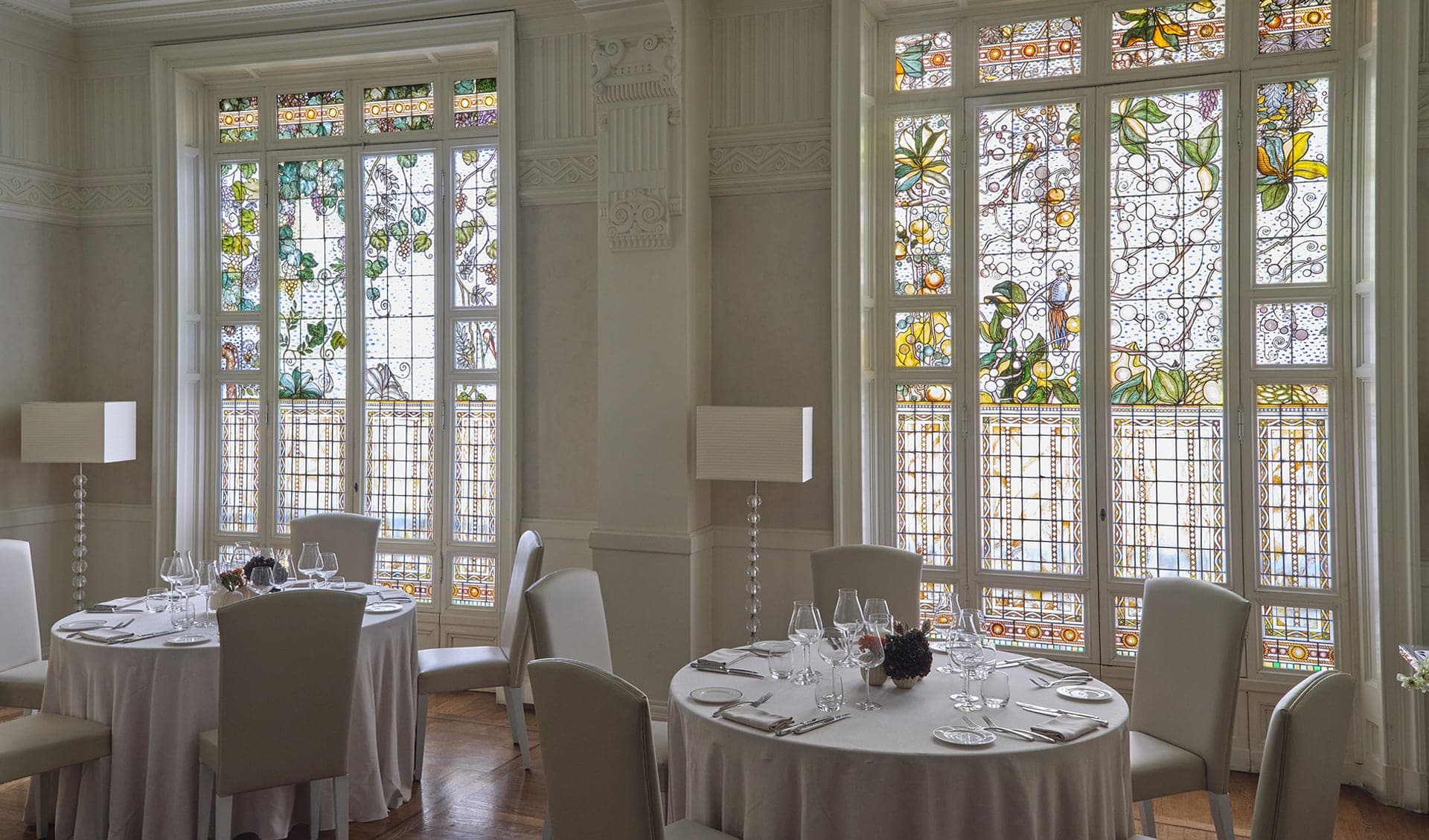 A dining room with round tables set for a meal, featuring large stained glass windows.
