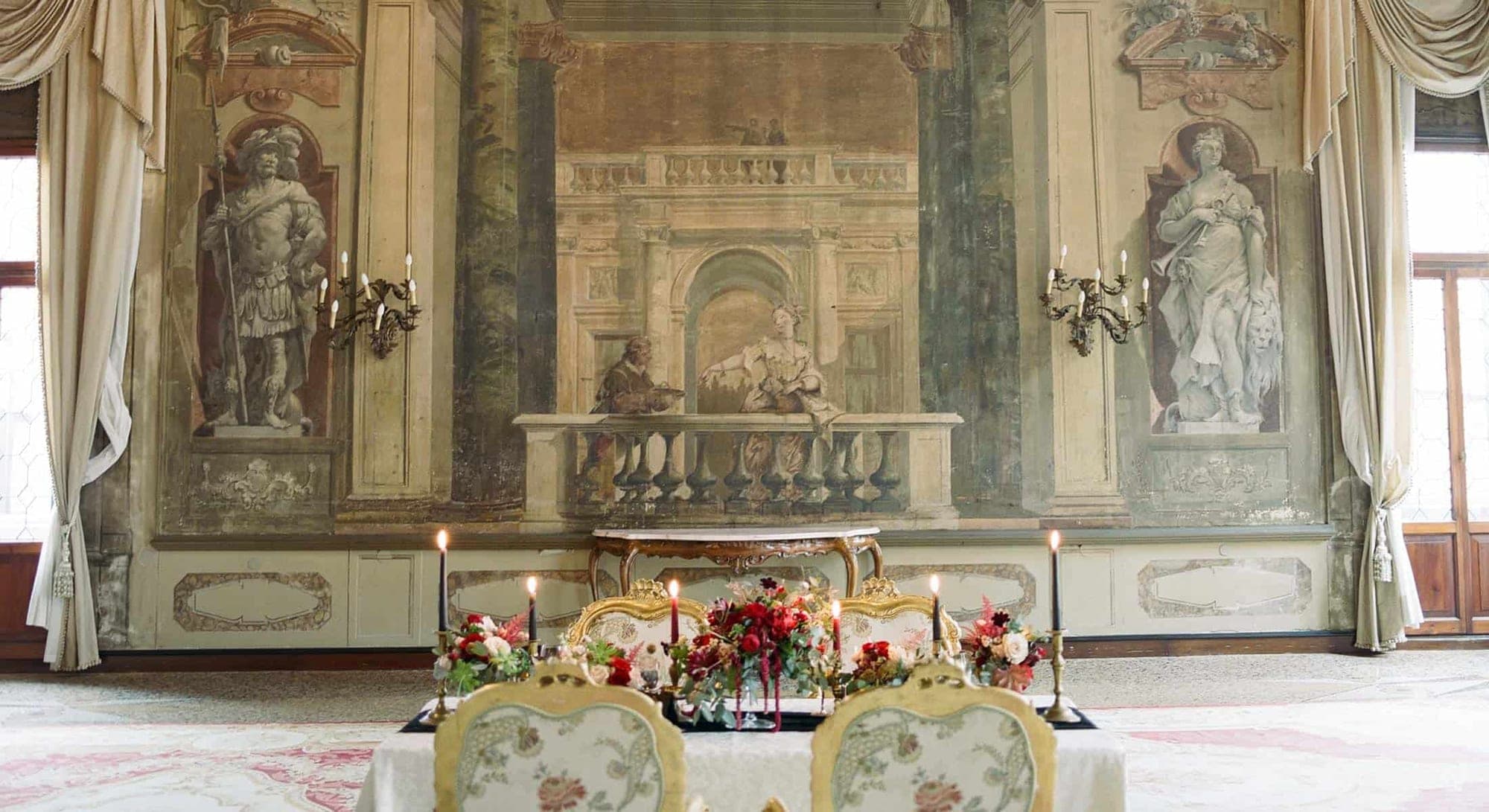A wedding reception table is set with floral arrangements and candles in a room with ornate frescoes.