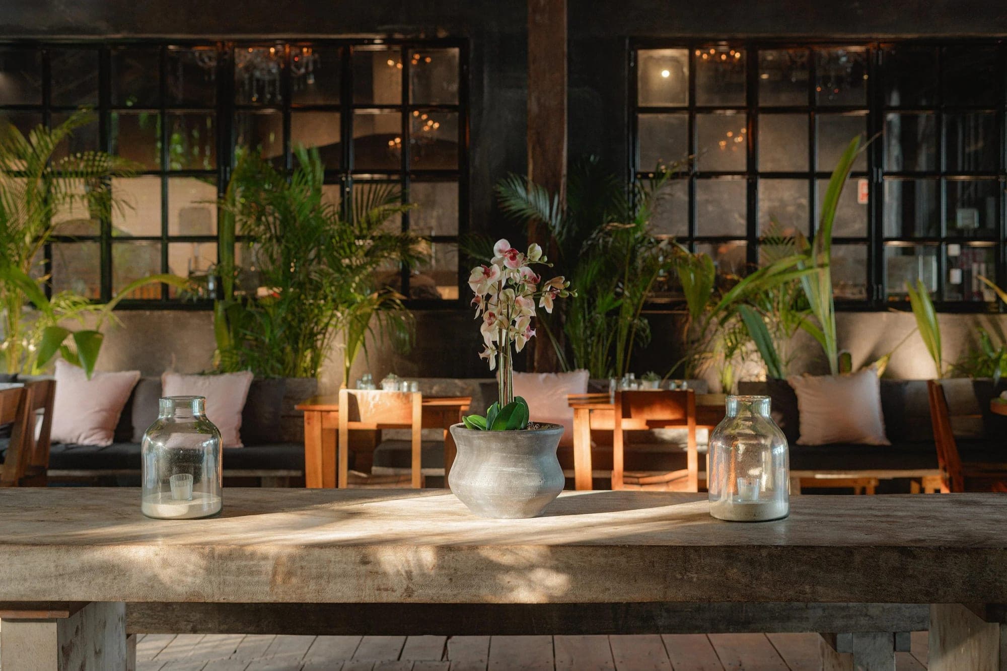 A wooden table with lanterns and a potted orchid in front of a seating area with large windows.