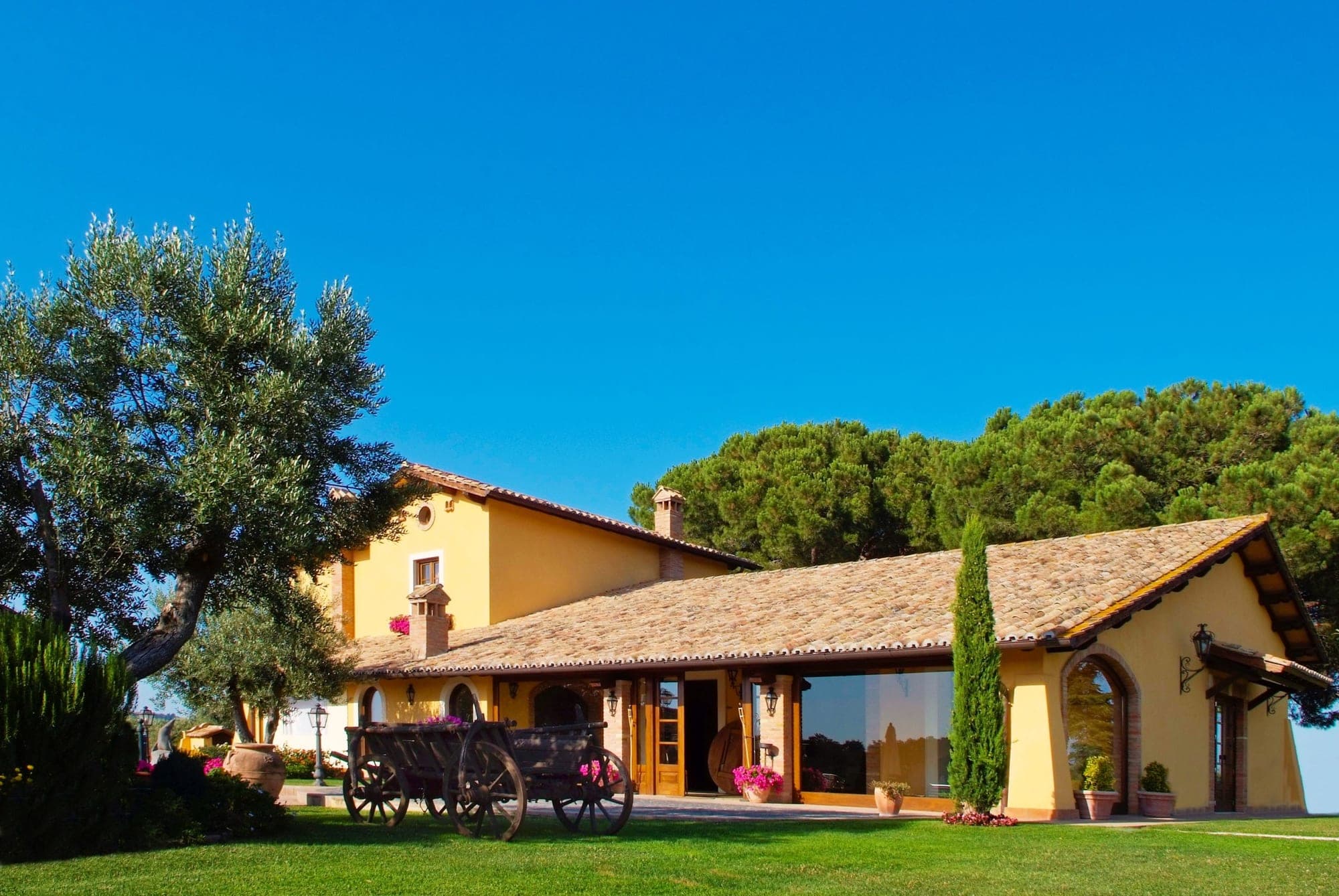 Yellow building with a tiled roof and large windows, set in a green lawn with trees.