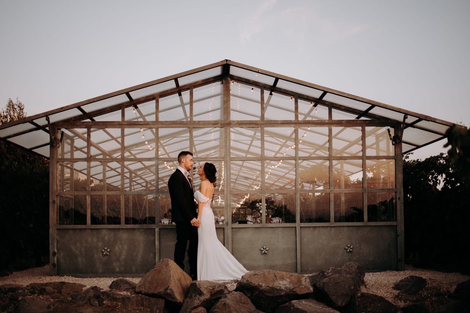 Couple in front of a greenhouse with string lights at dusk.