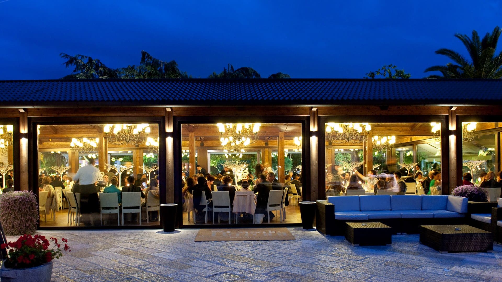 A wedding reception is underway inside a glass-walled building, viewed from an outdoor patio.