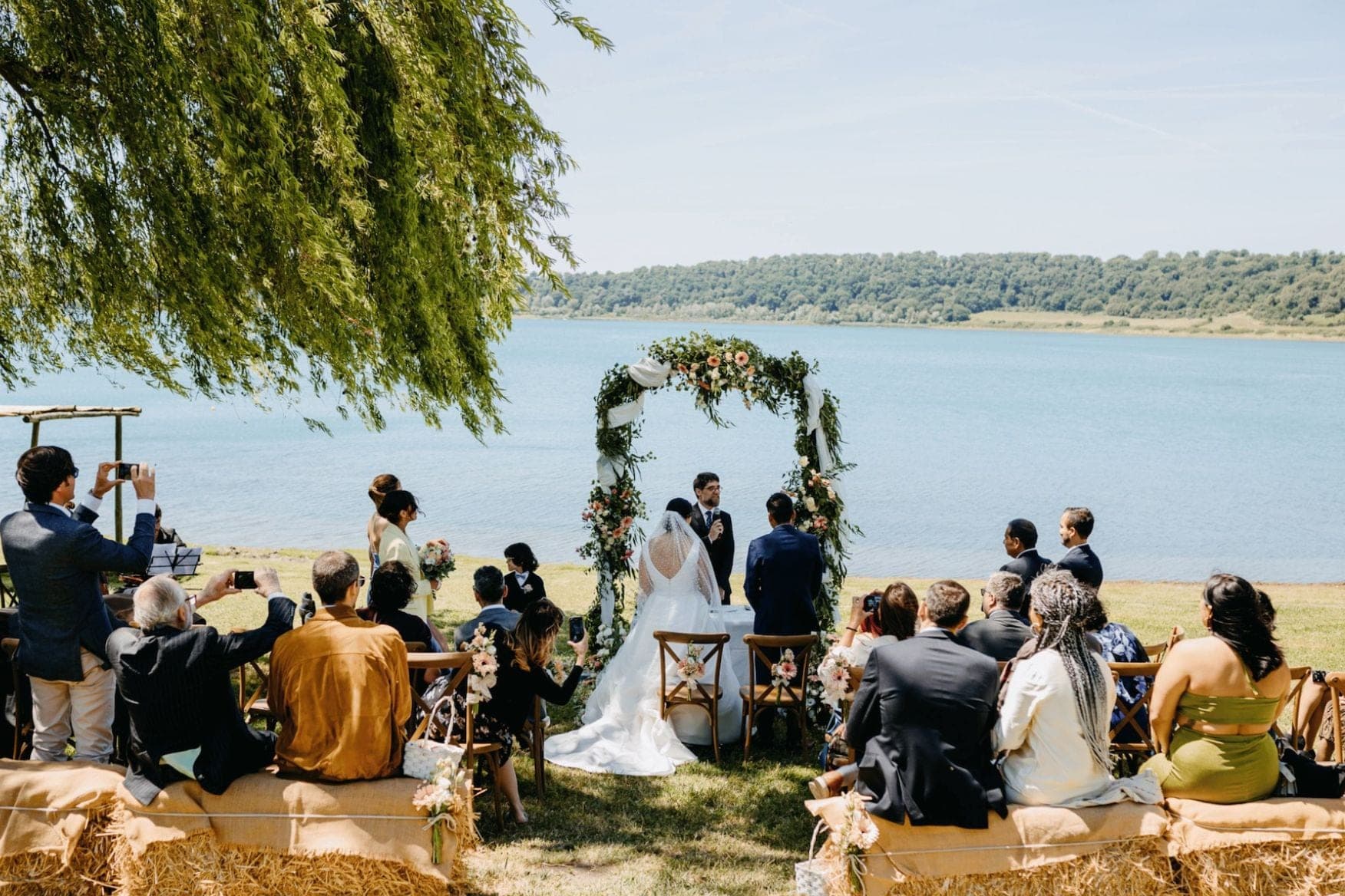 Outdoor wedding ceremony by a lake with guests seated on hay bales.