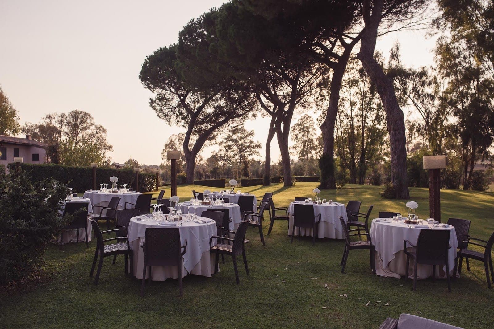 Tables set for a reception in a garden with trees and a building in the background.