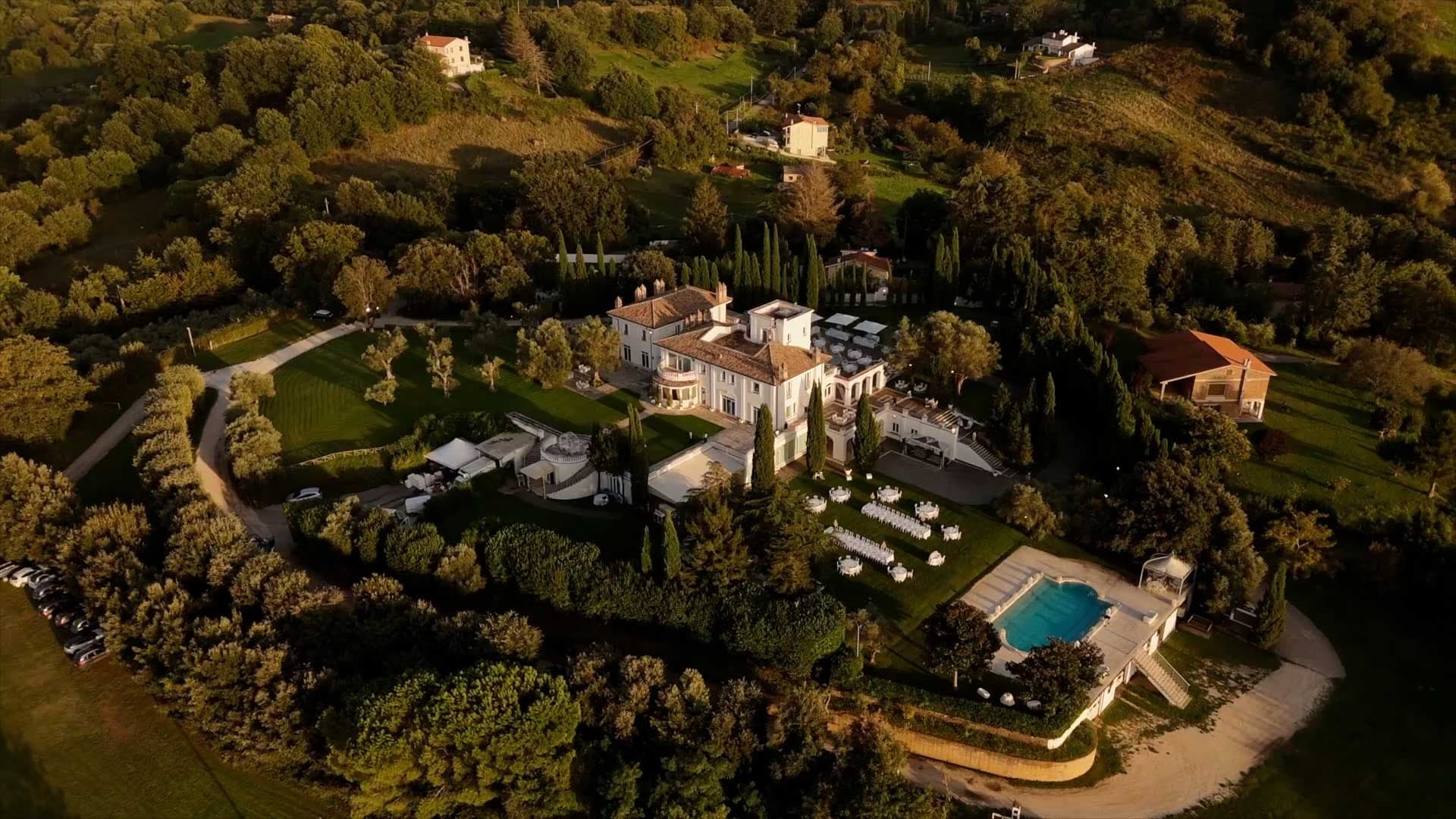 Aerial view of a large white villa with a swimming pool and reception tables set up on the lawn.