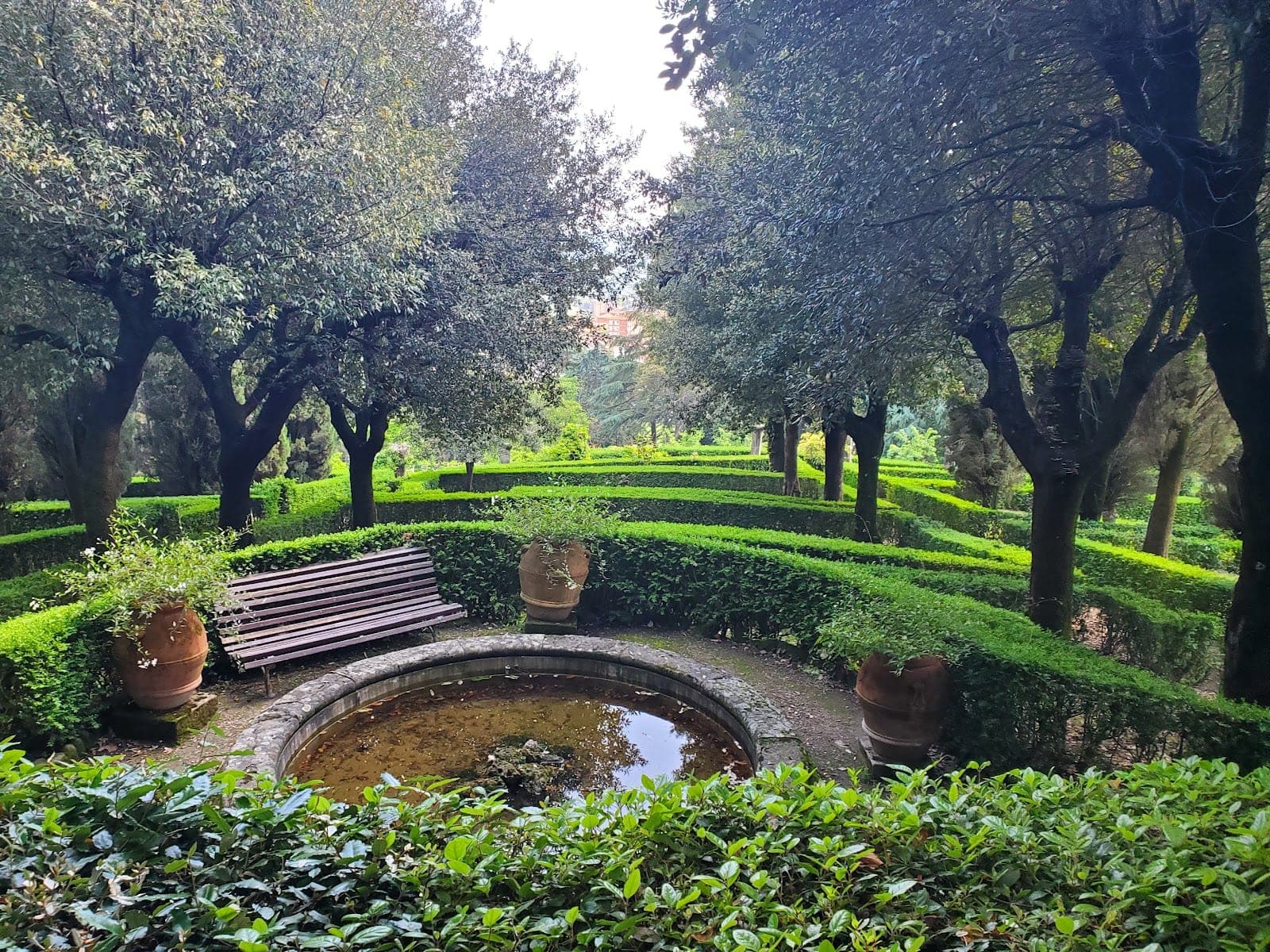 A formal garden with manicured hedges, trees, a bench, and a small fountain.