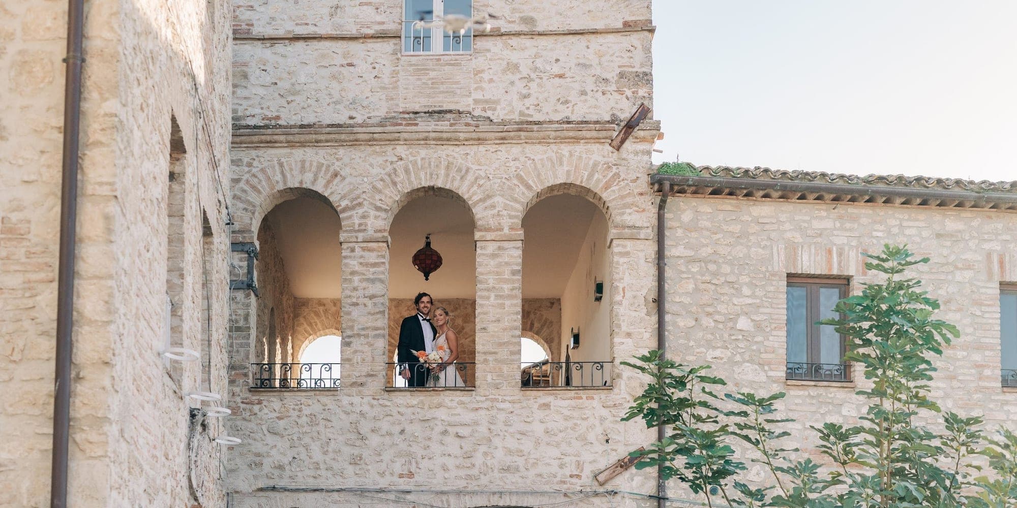 A bride and groom stand on a balcony of a stone building.