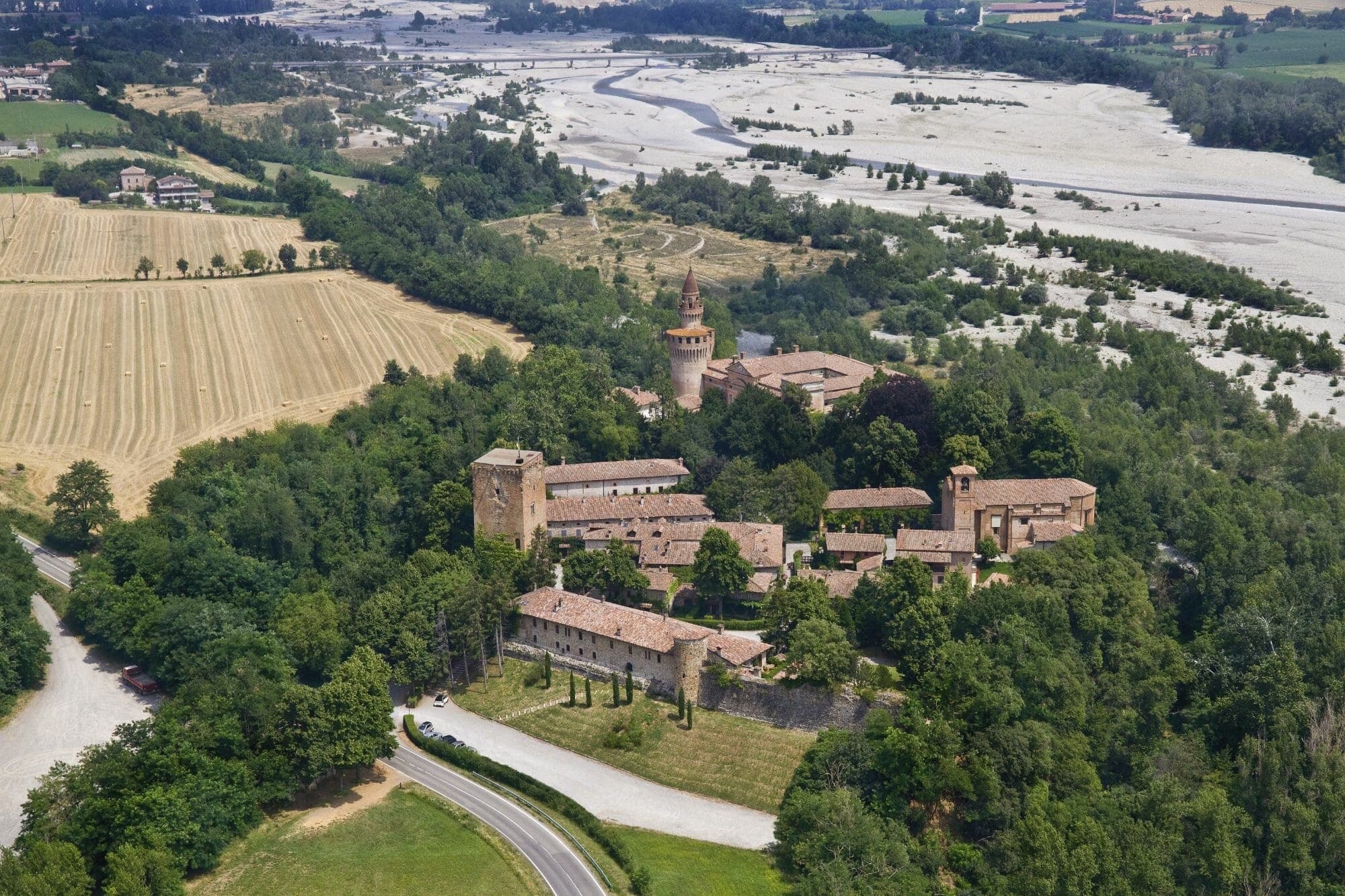 Aerial view of Castello di Rivalta surrounded by trees and fields, with a river in the distance.