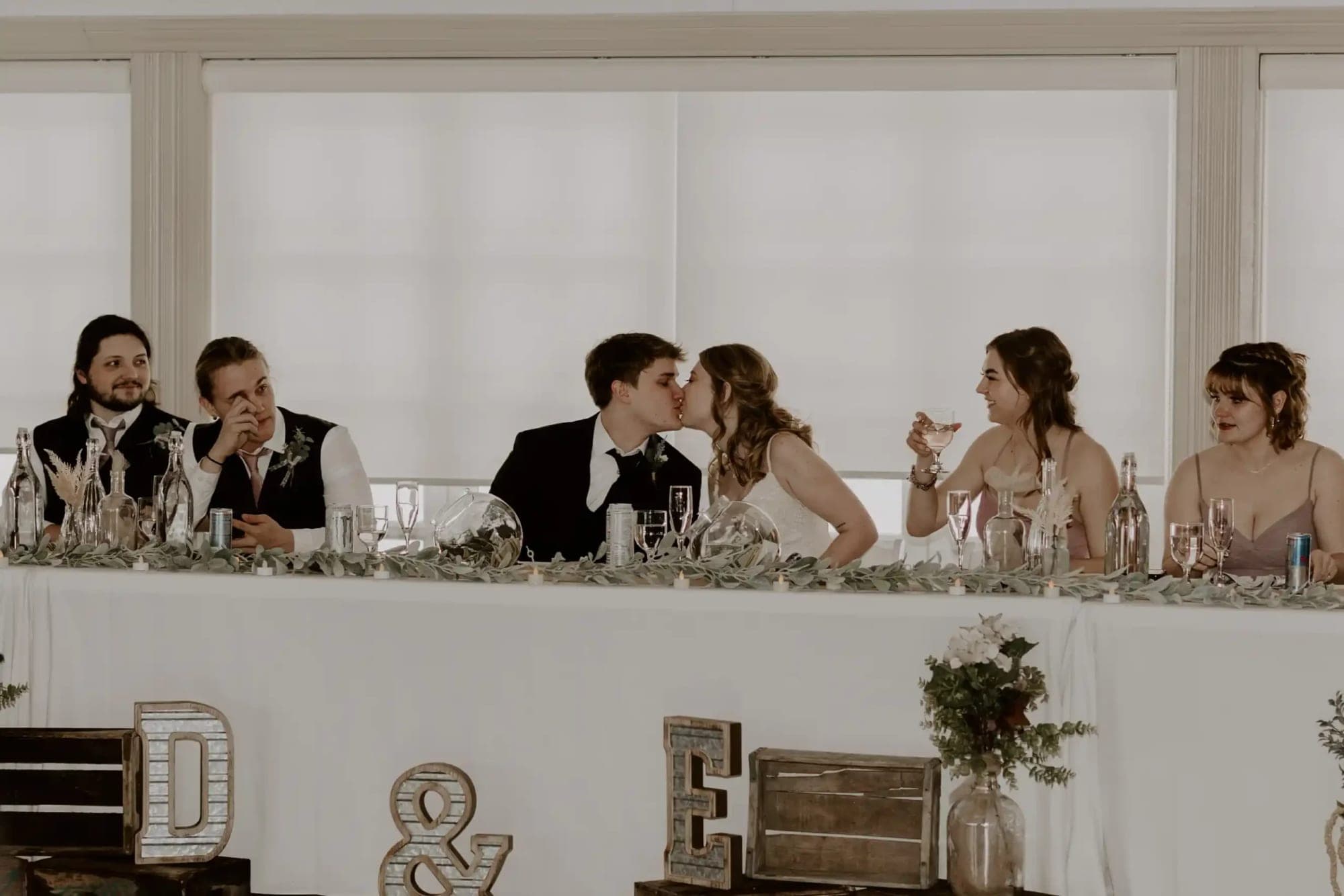 Wedding party at a reception table, with the bride and groom kissing.