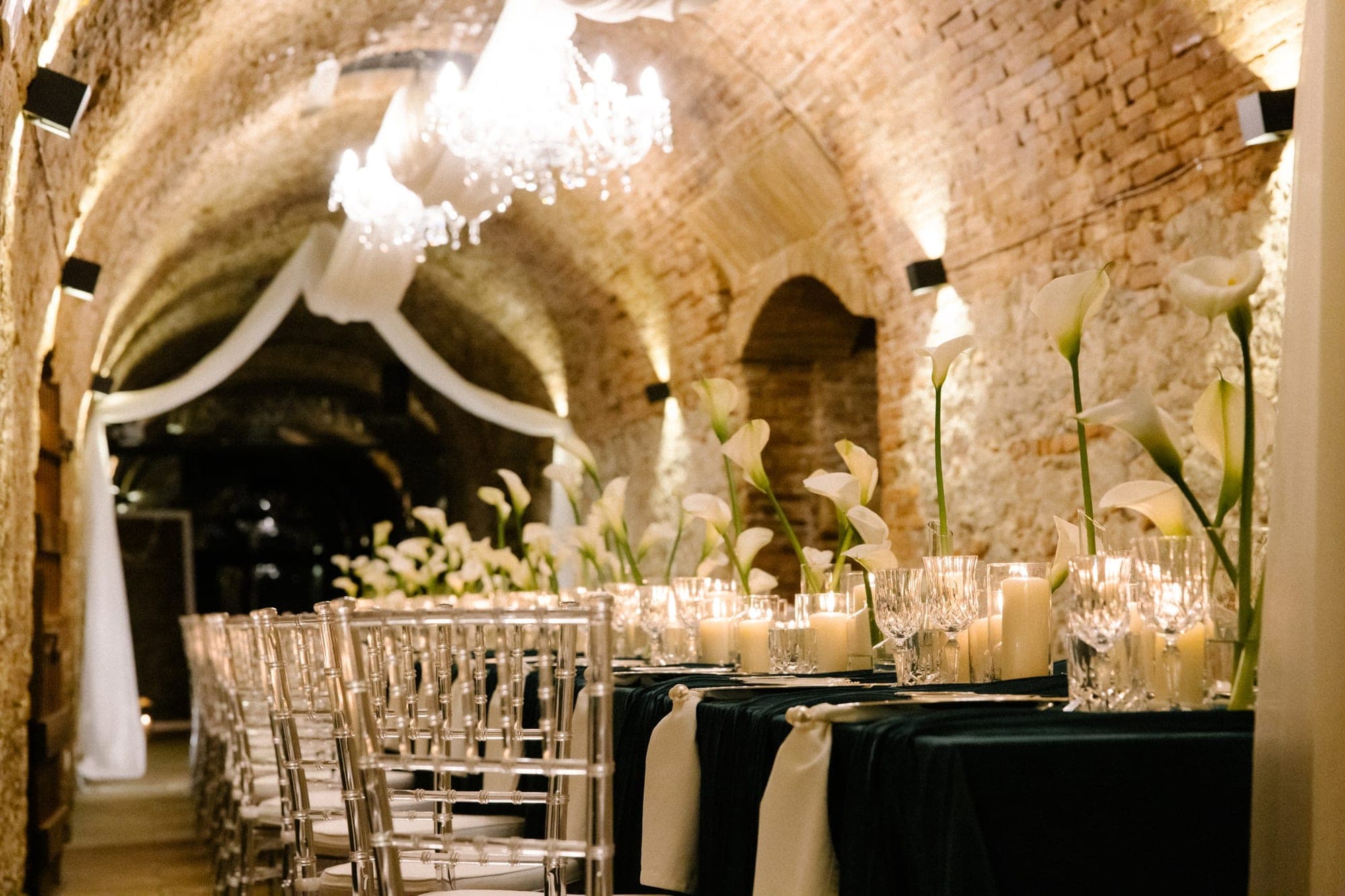 A long table set for a wedding reception in a stone vaulted room with chandeliers.