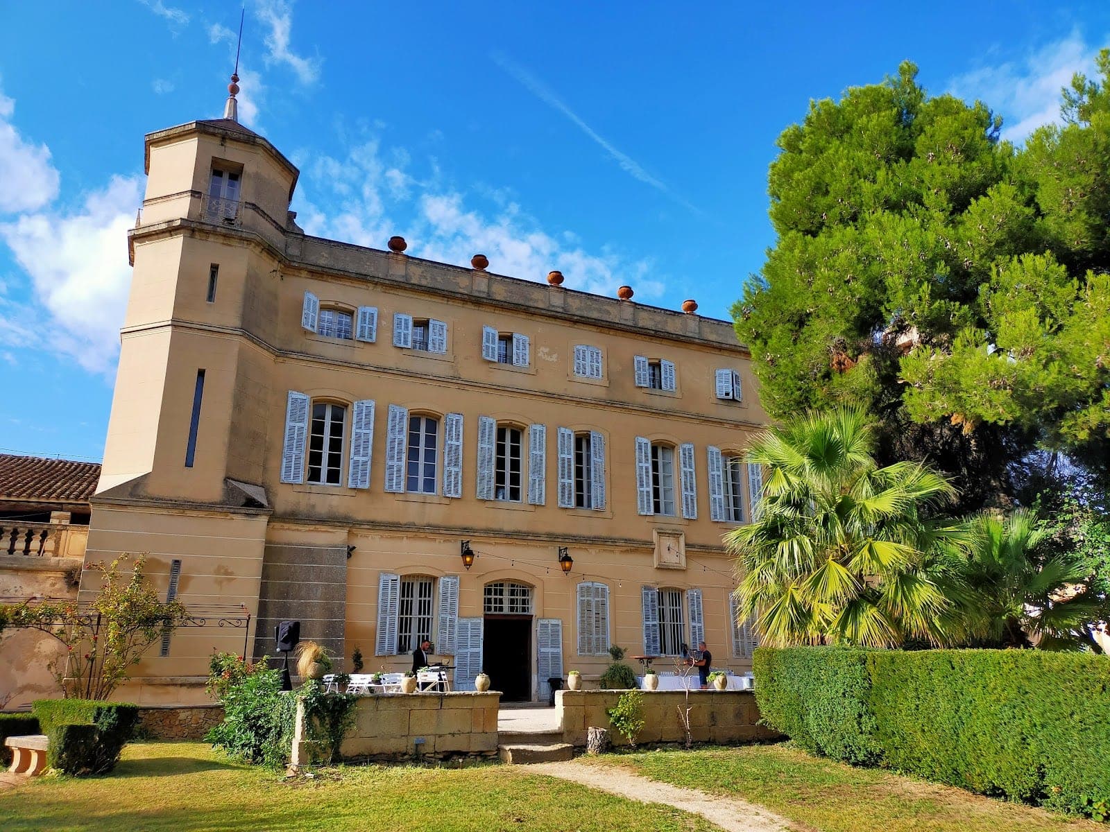 Exterior of a pale yellow building with blue shutters and a tower, set in green grounds.
