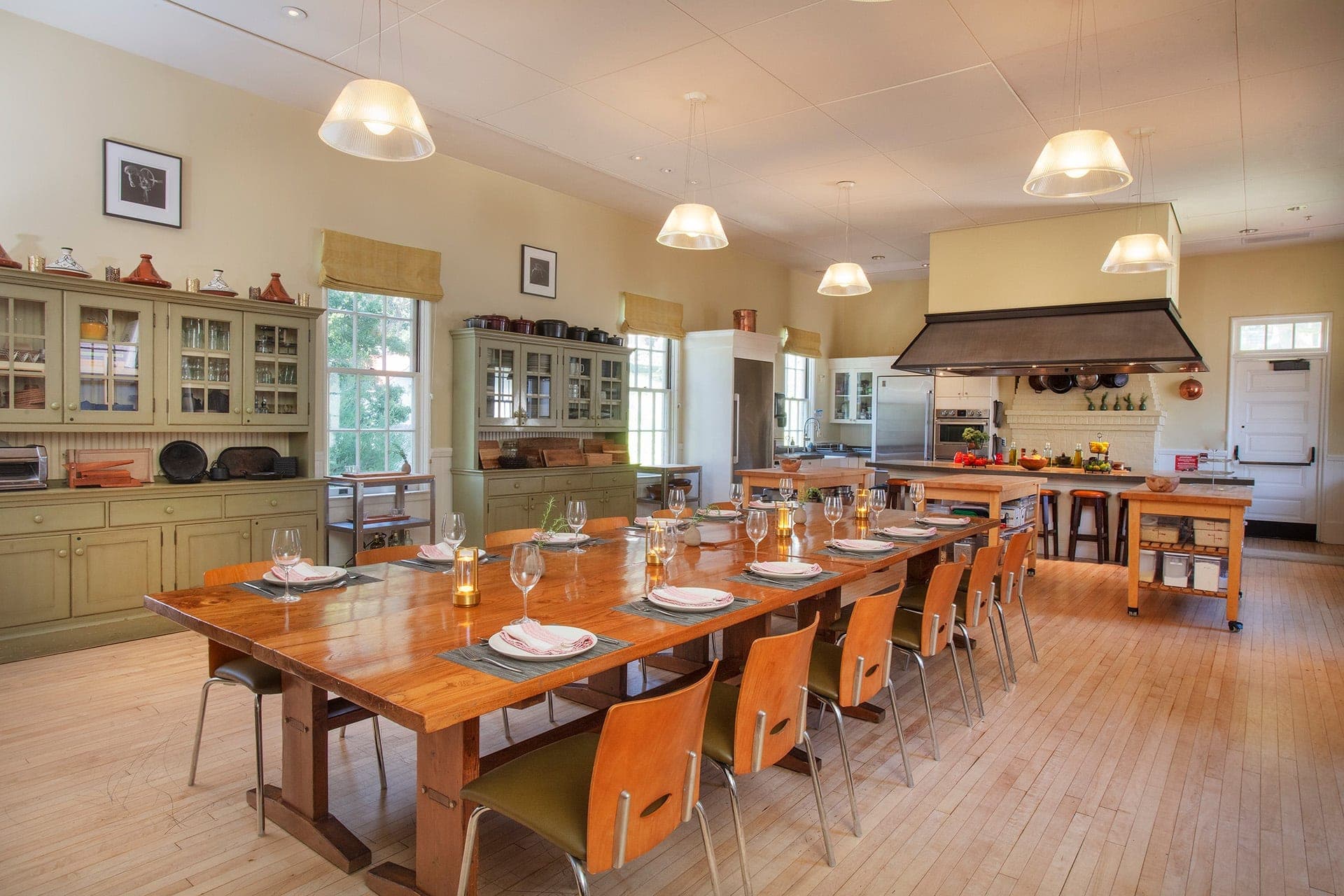 A long wooden dining table is set with plates and glasses in a room with kitchen cabinets and a large stove.