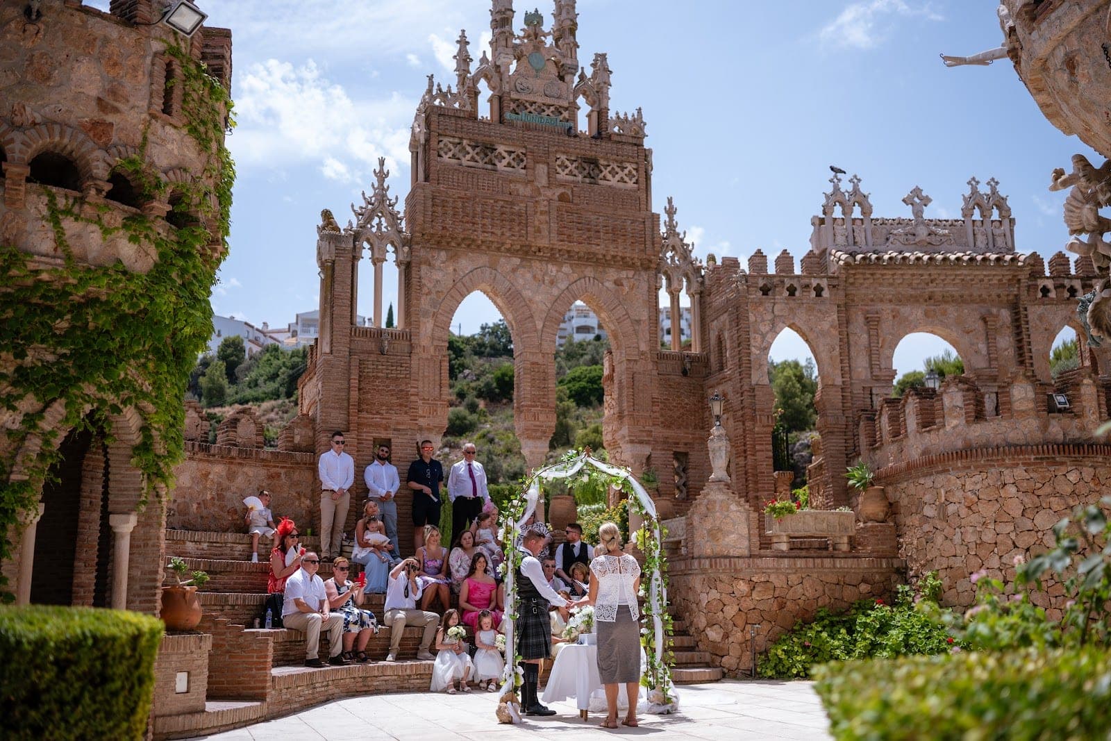 Wedding ceremony in front of a stone castle with guests seated on steps.