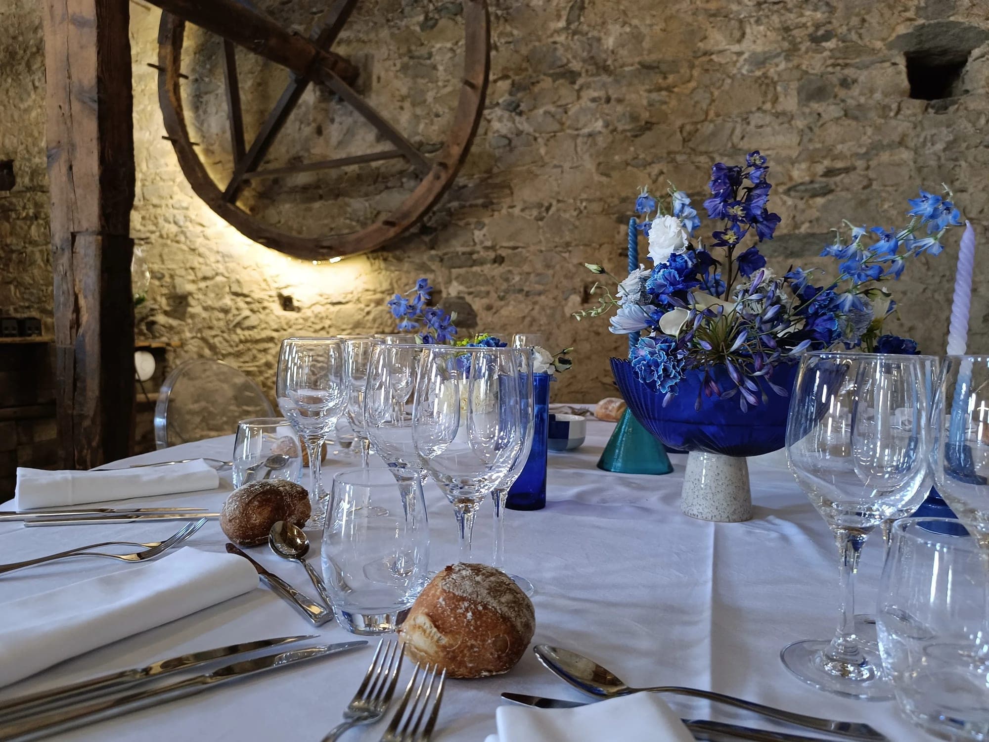 A wedding reception table is set with white linens, glassware, cutlery, bread rolls, and a blue vase with blue and white flowers.
