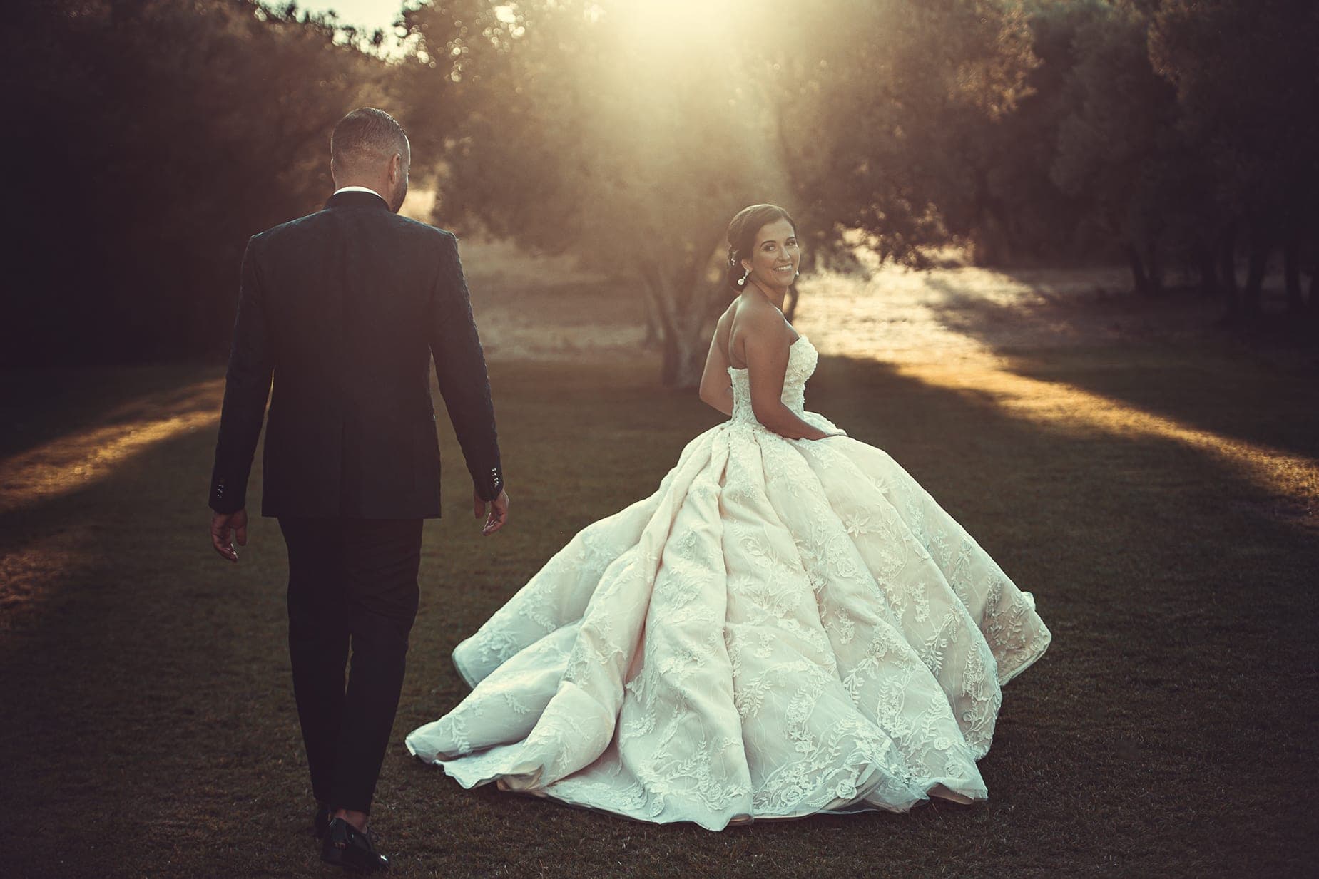 Bride and groom walking on grass with sunbeams through trees