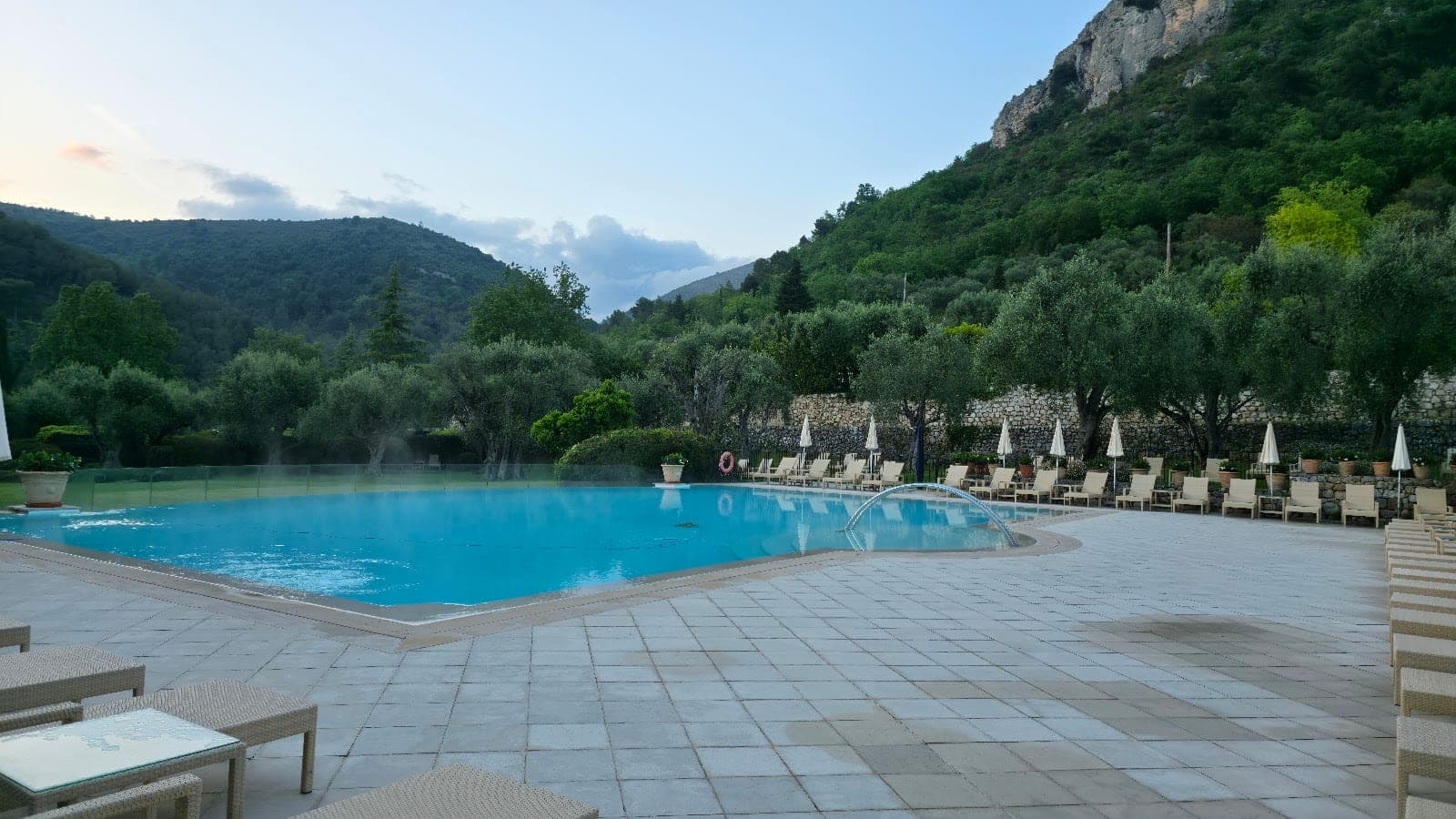 Swimming pool area with loungers and umbrellas, set against green hills.