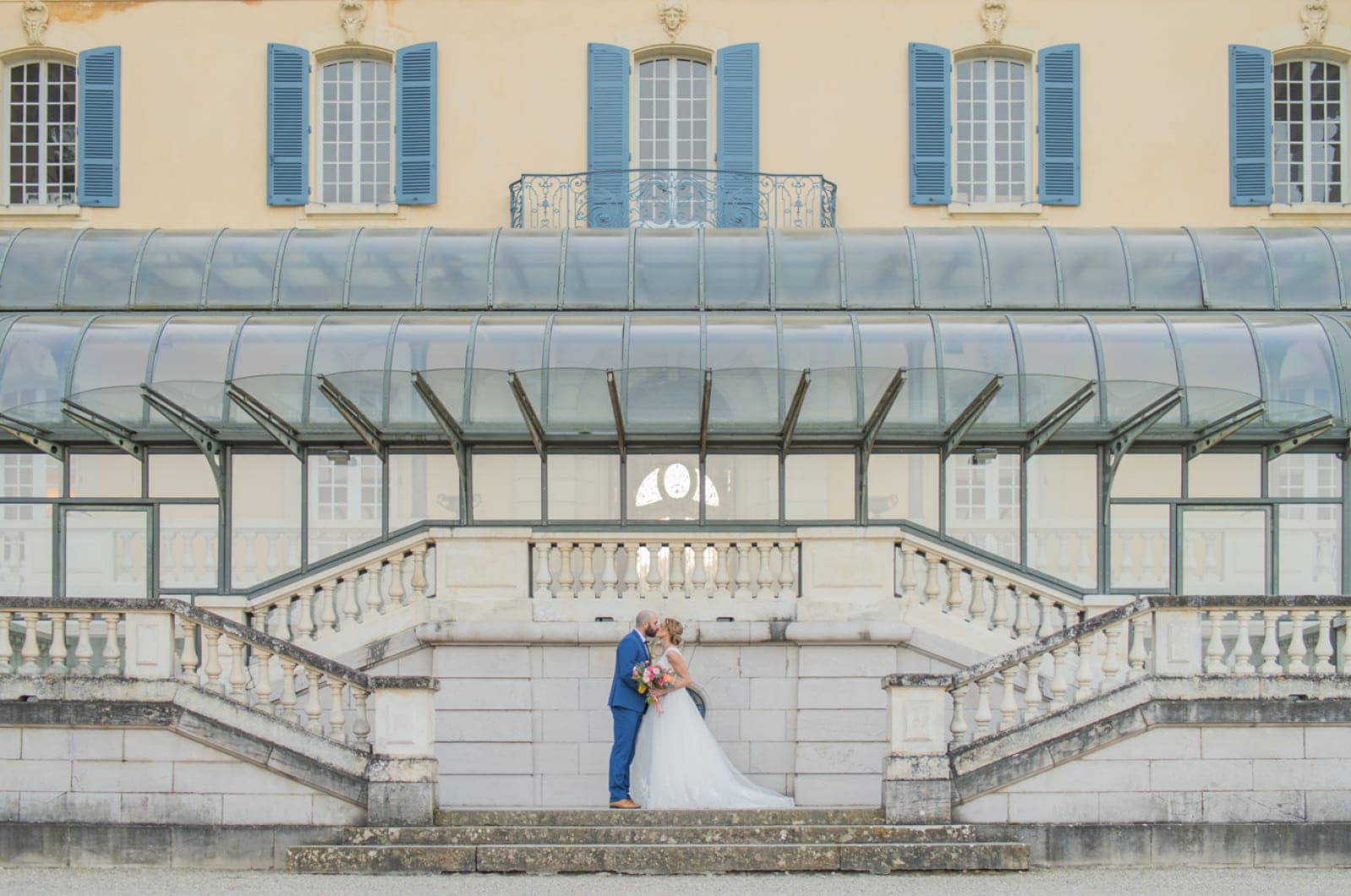 A bride and groom kiss on a stone staircase in front of a yellow chateau with blue shutters.