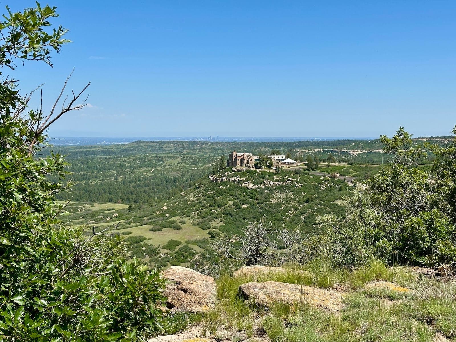A castle sits on a hill overlooking a valley with a distant city skyline under a clear blue sky.