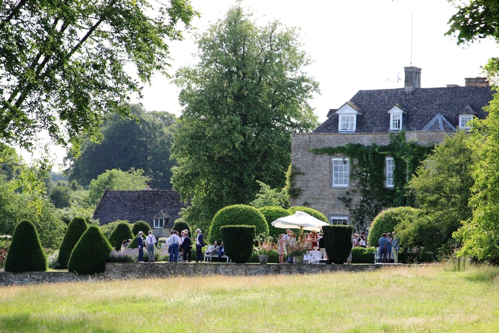 Manor house with gardens and people gathered outside on a sunny day.