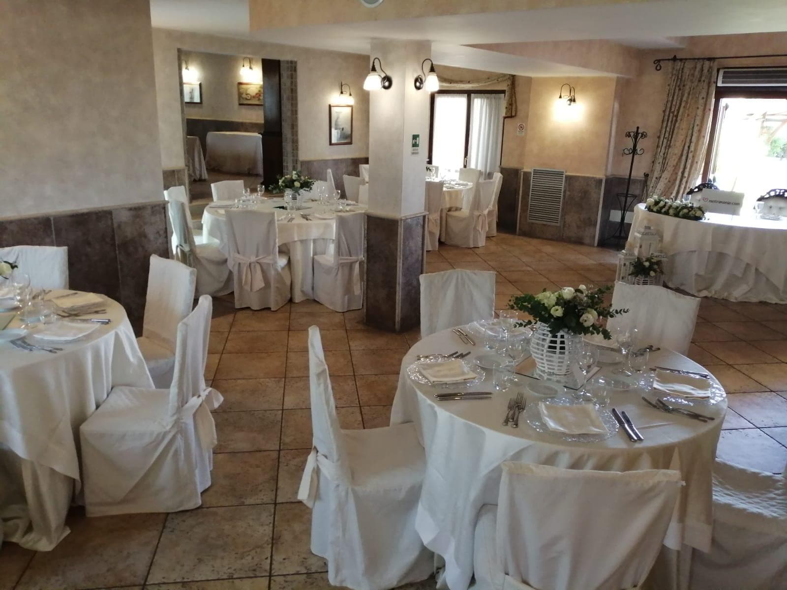 A wedding reception dining room with round tables covered in white tablecloths and chairs with white covers.