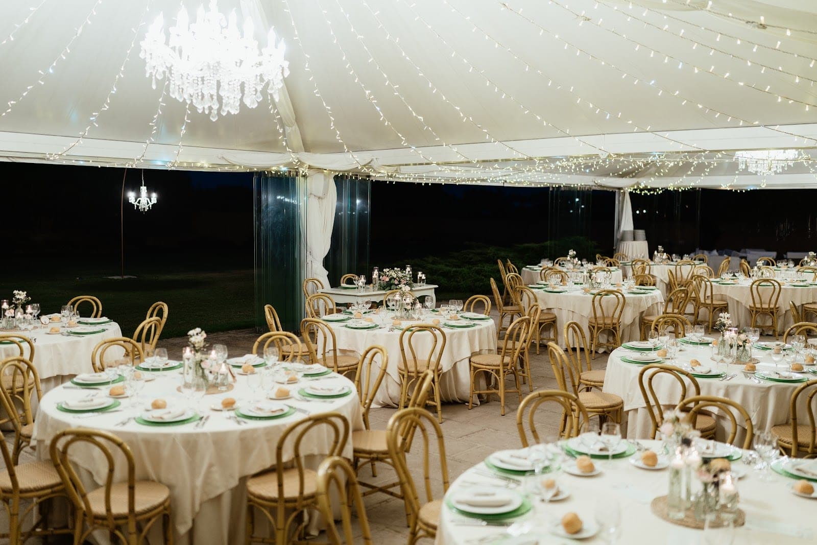A reception tent with round tables set for dinner, lit by fairy lights and chandeliers.
