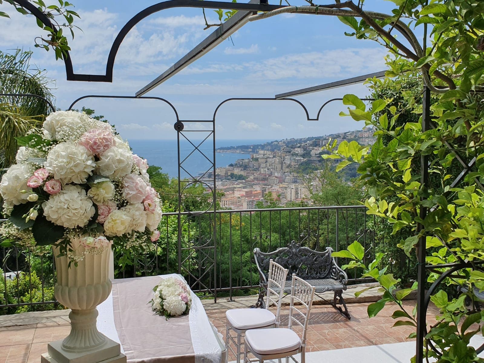 Terrace set for a wedding ceremony with floral arrangements and a view of the city and sea.