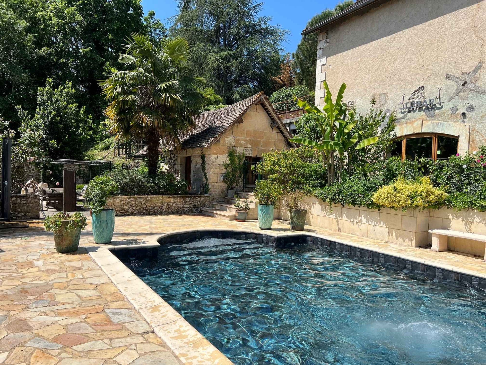 A swimming pool with blue water and a stone patio area next to a stone building and garden.